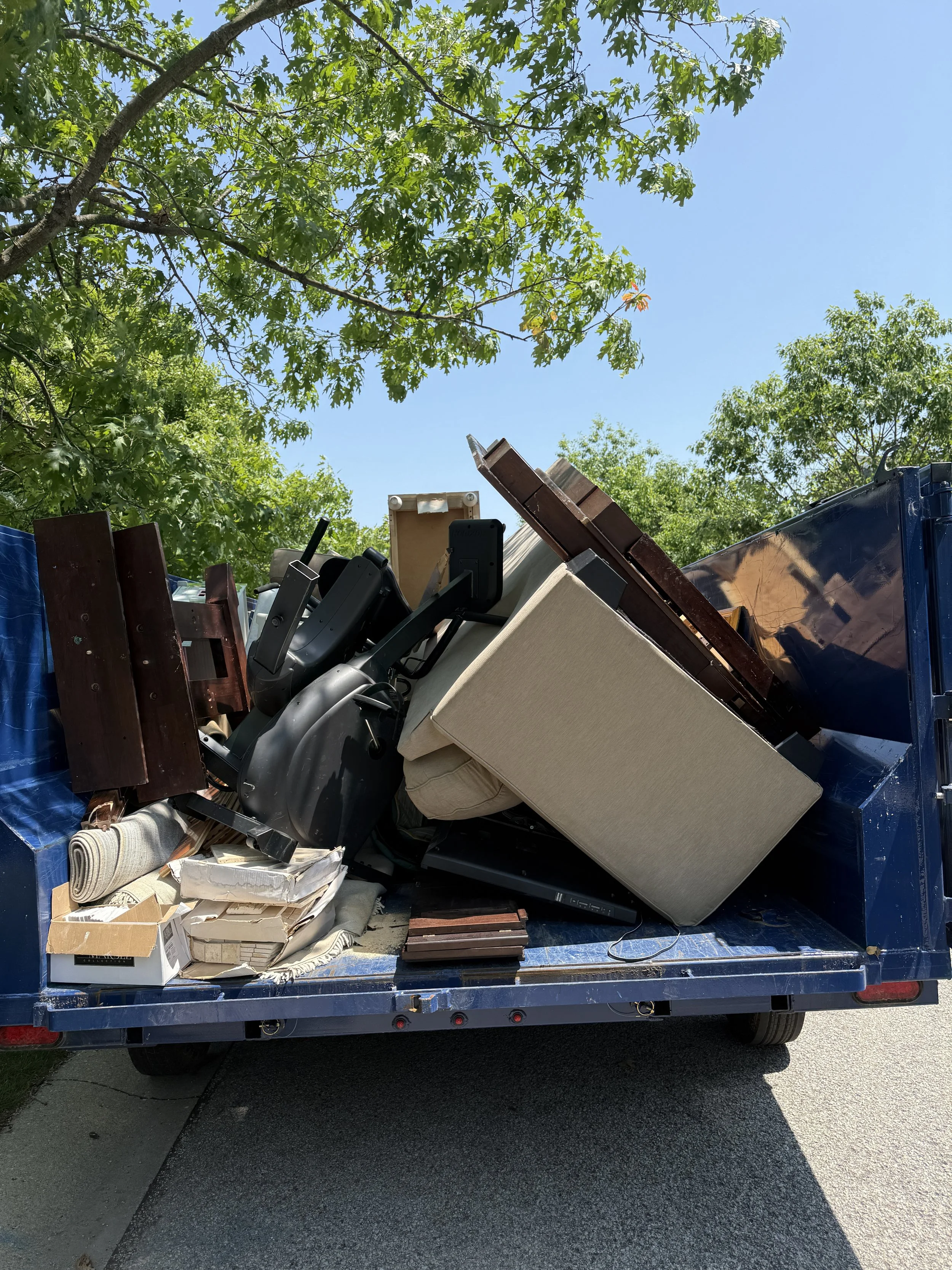 Clean outdoor blue truck loaded with furniture and household items, parked on a residential street with green trees and clear blue sky in the background.
