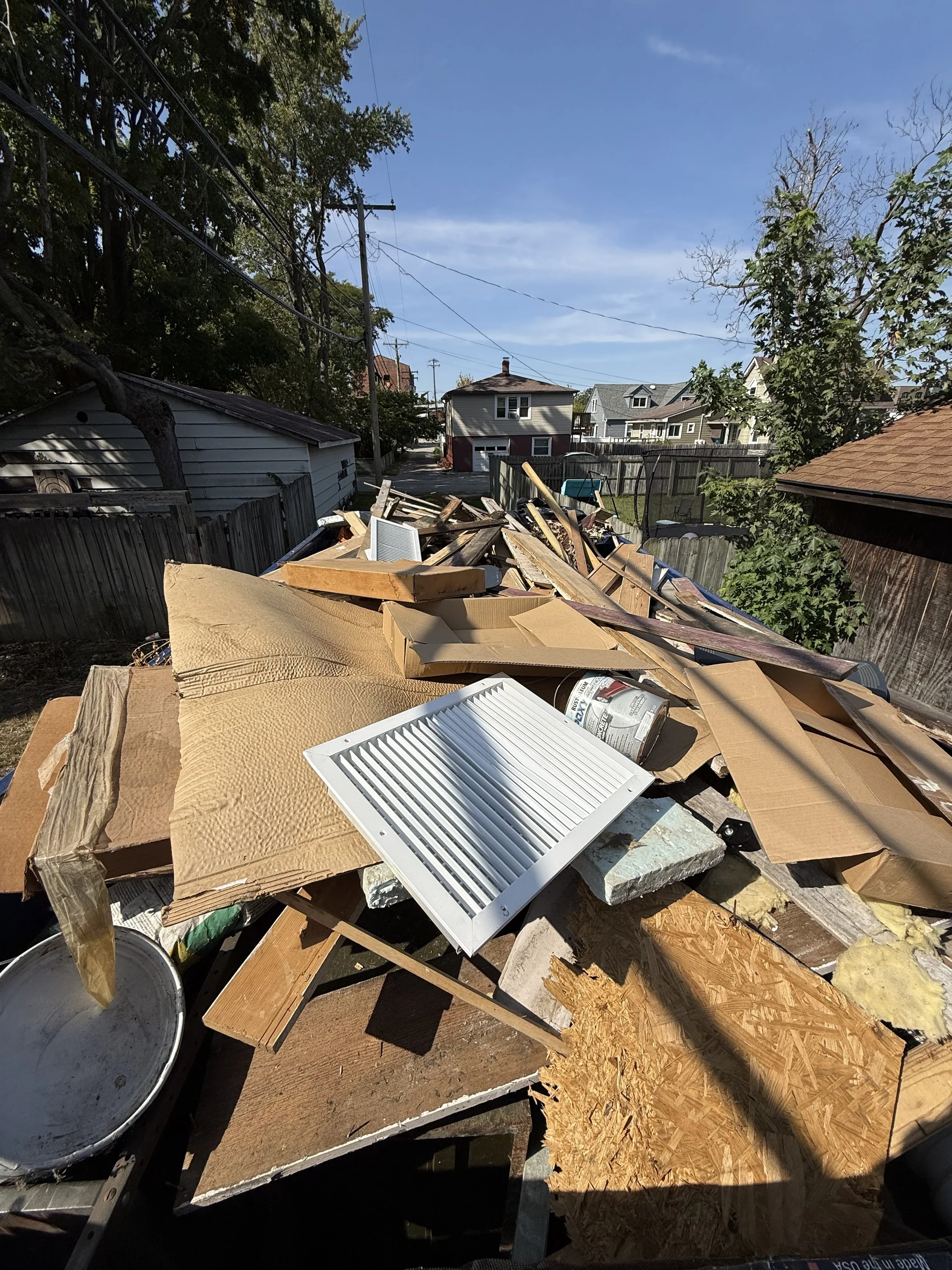 Piles of discarded household materials and debris, including wood, cardboard, and metal, outdoors in a backyard.
