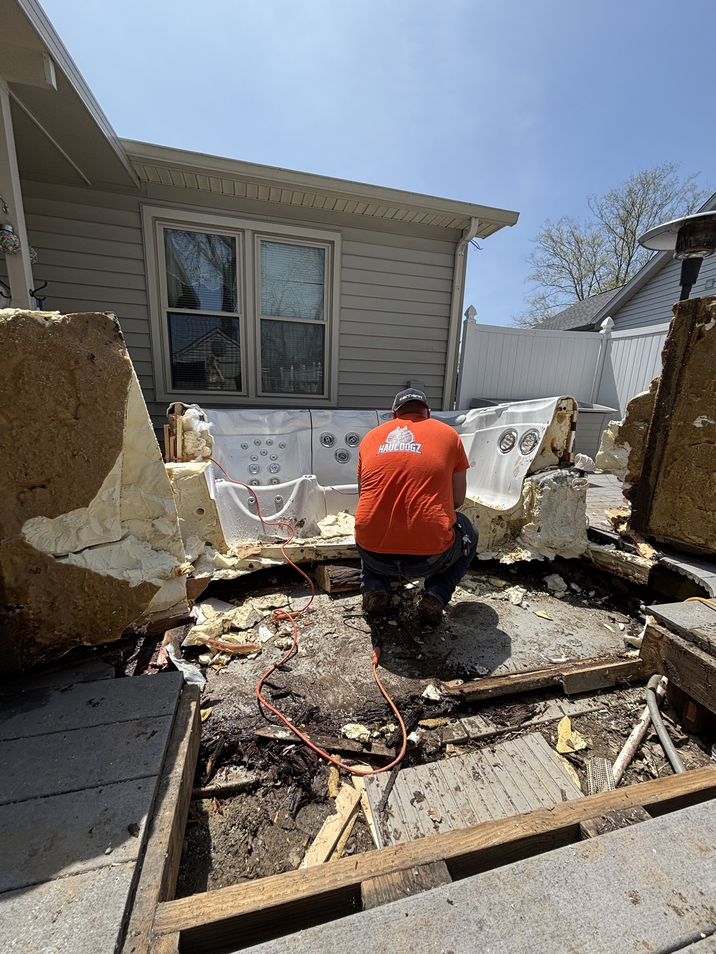 Worker kneeling in a backyard removing a hot tub with saws and tools, surrounded by debris, with a house and a white fence in the background.