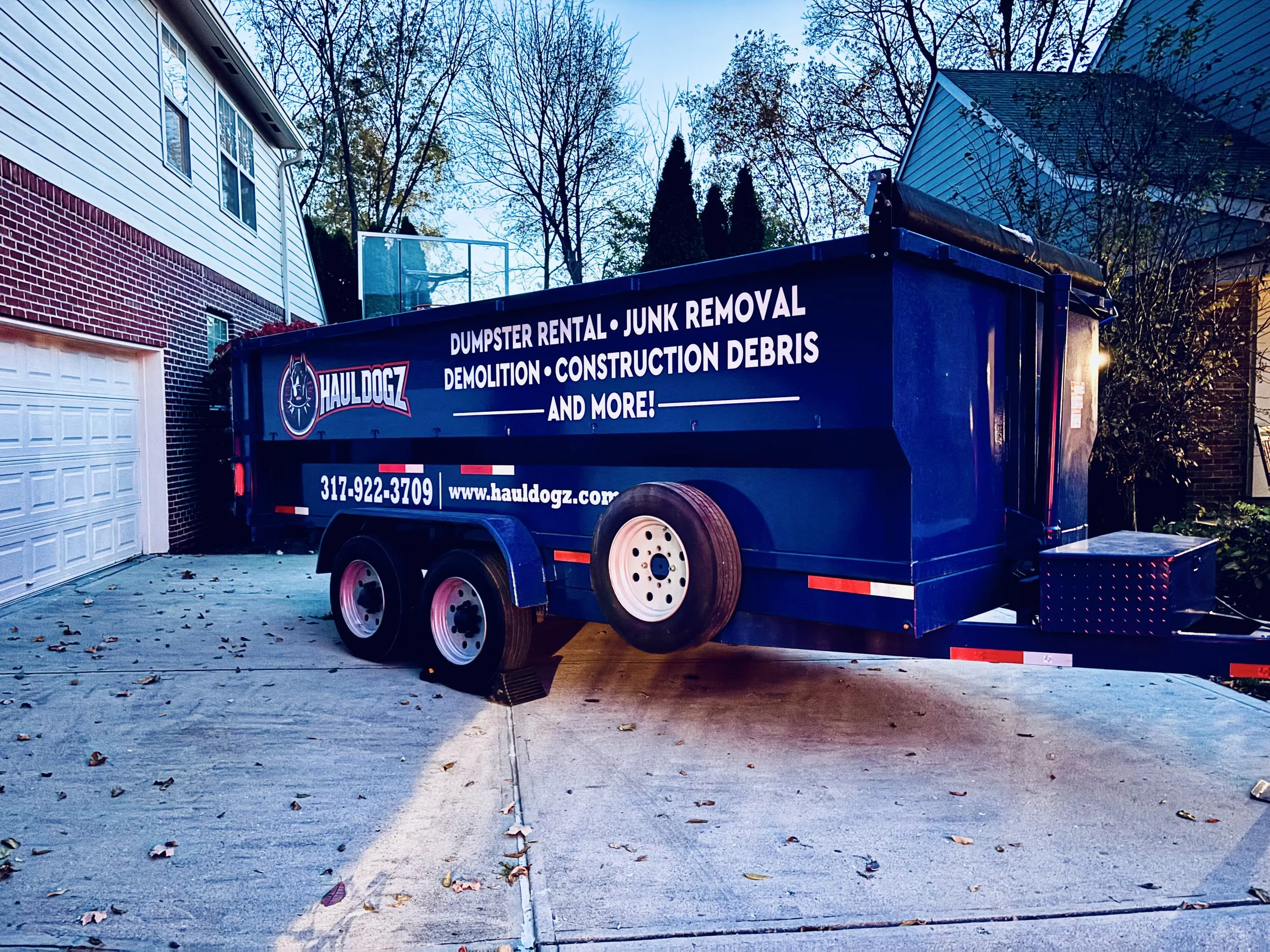 A blue trailer parked in a driveway with advertising for Hauldogz, a junk removal and demolition service, with contact information and website. There is a house and trees in the background.