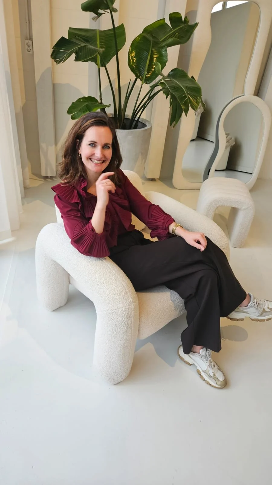 Vrouw zit op een moderne beige stoel, lachend, in een interieur met grote groene plant en spiegels.