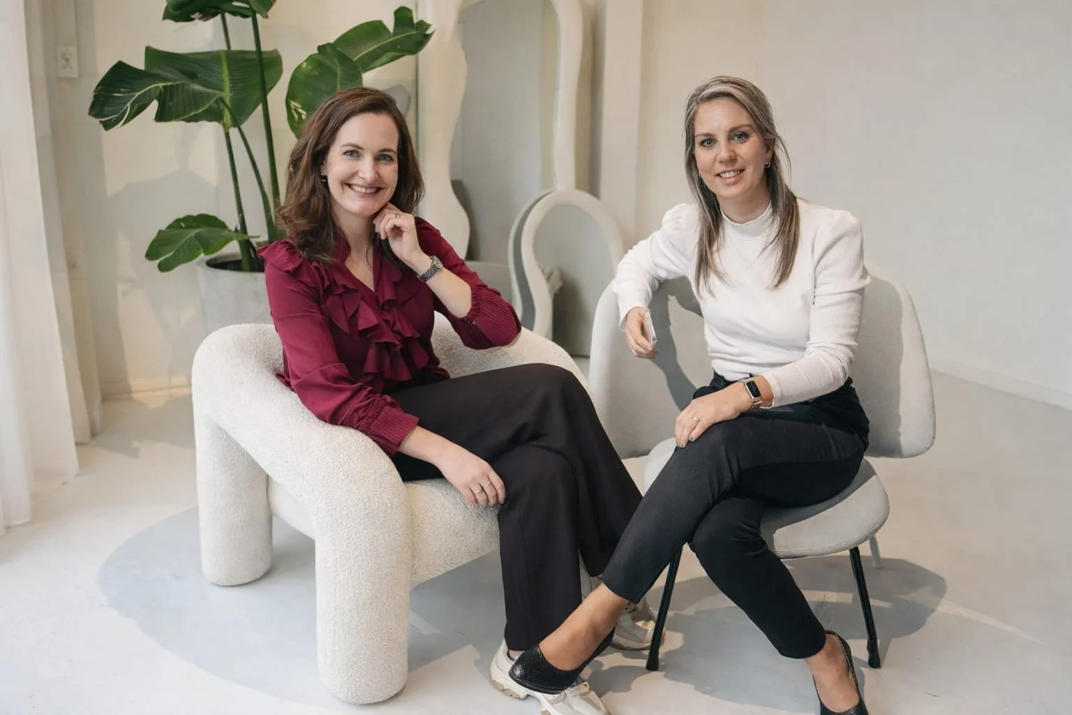 Twee vrouwen zitten ontspannen in een lichte, minimalistische kamer met een grote groene plant op de achtergrond.