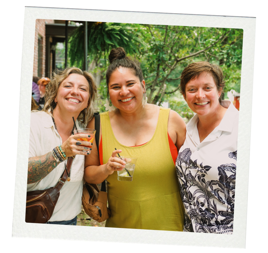 Three women smiling outdoors at a social gathering, holding drinks, with trees and a building in the background.
