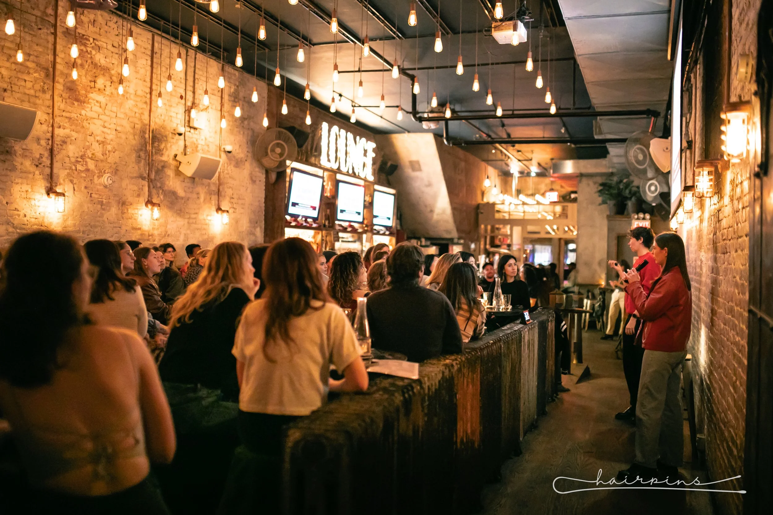 A group of people sitting and watching a presentation in a dimly lit, rustic bar or lounge with hanging string lights and exposed brick walls.