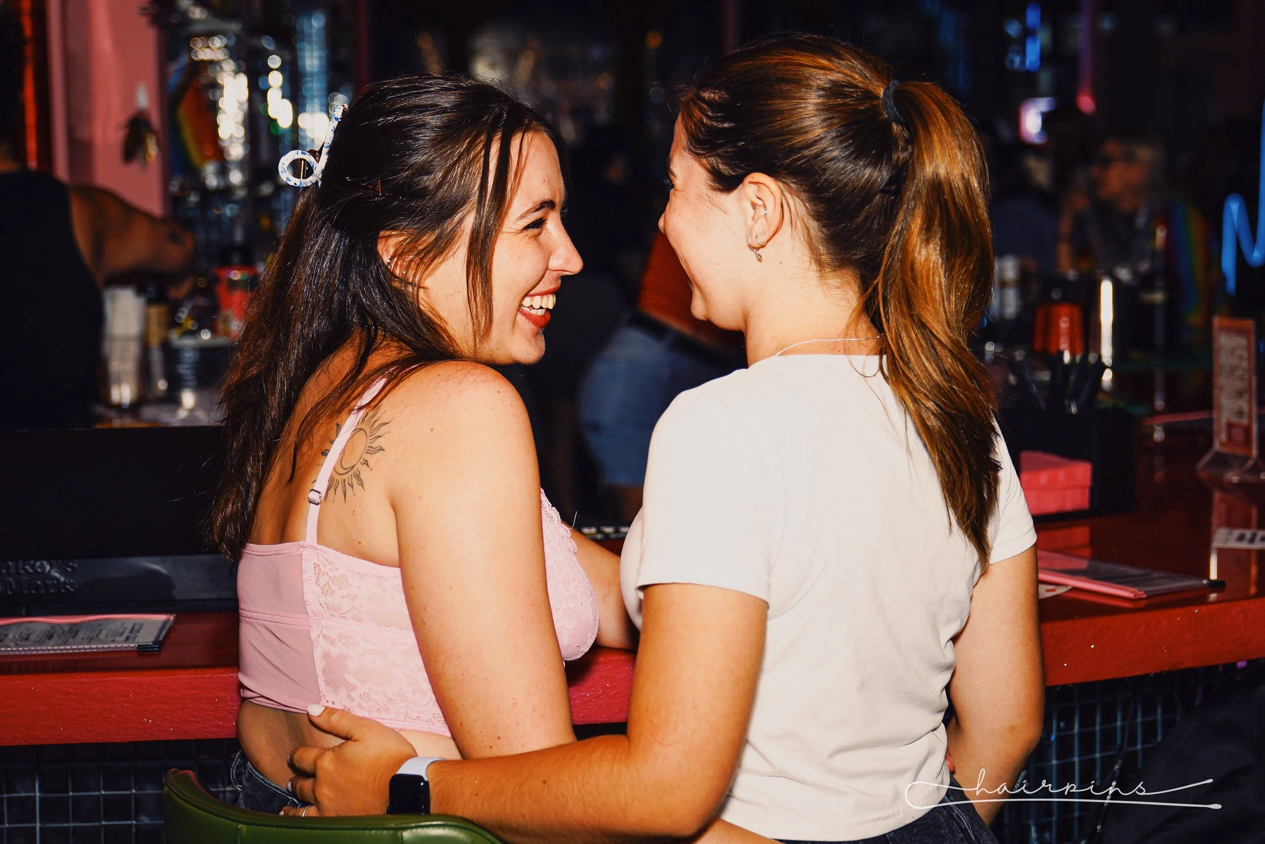 Two women smiling and enjoying a moment together at a bar or club, with a festive, colorful background.