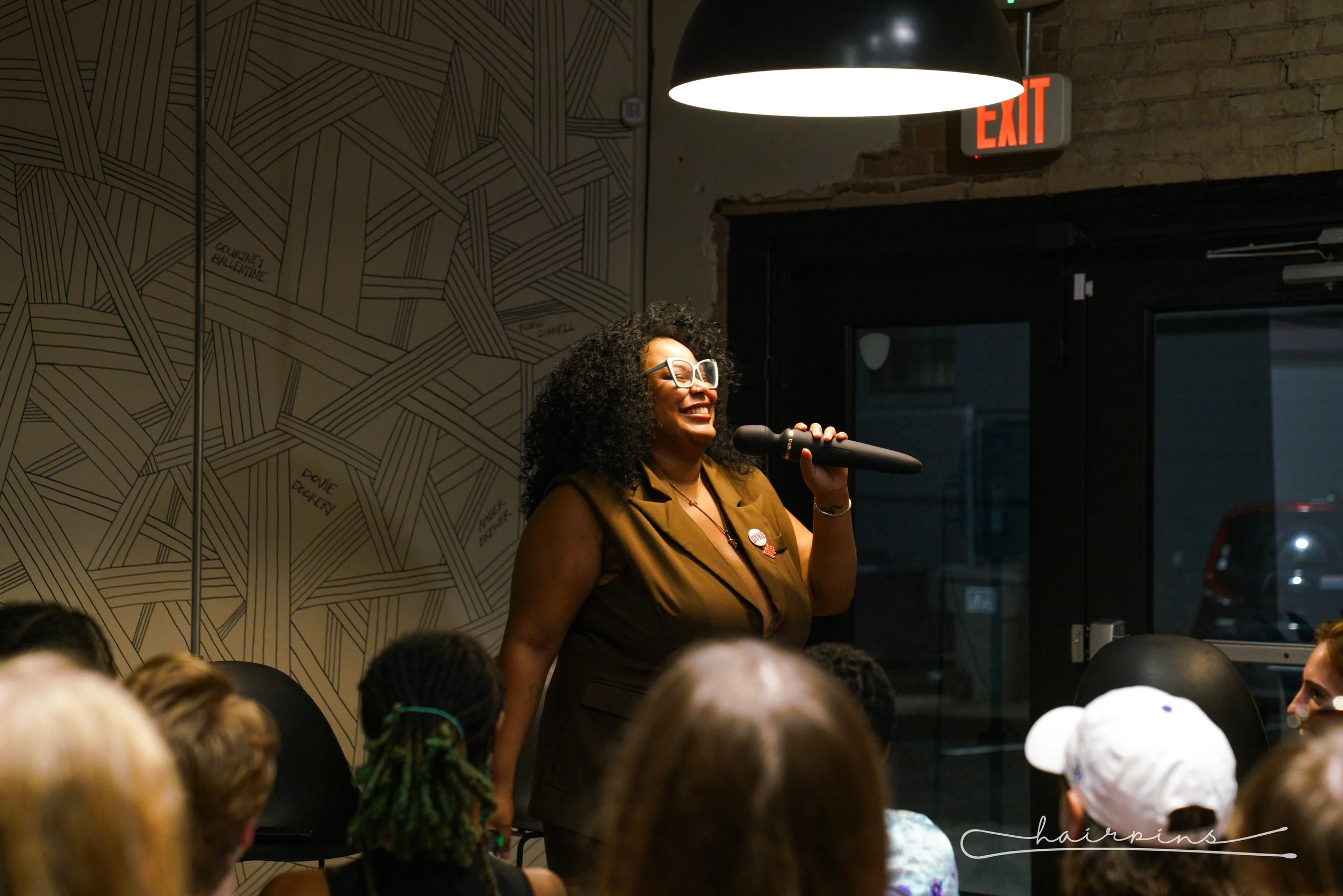 A woman with curly hair, glasses, and a brown vest is speaking into a microphone at a gathering. She is smiling and standing in front of an audience in a room with a patterned wall and an exit sign above the door.
