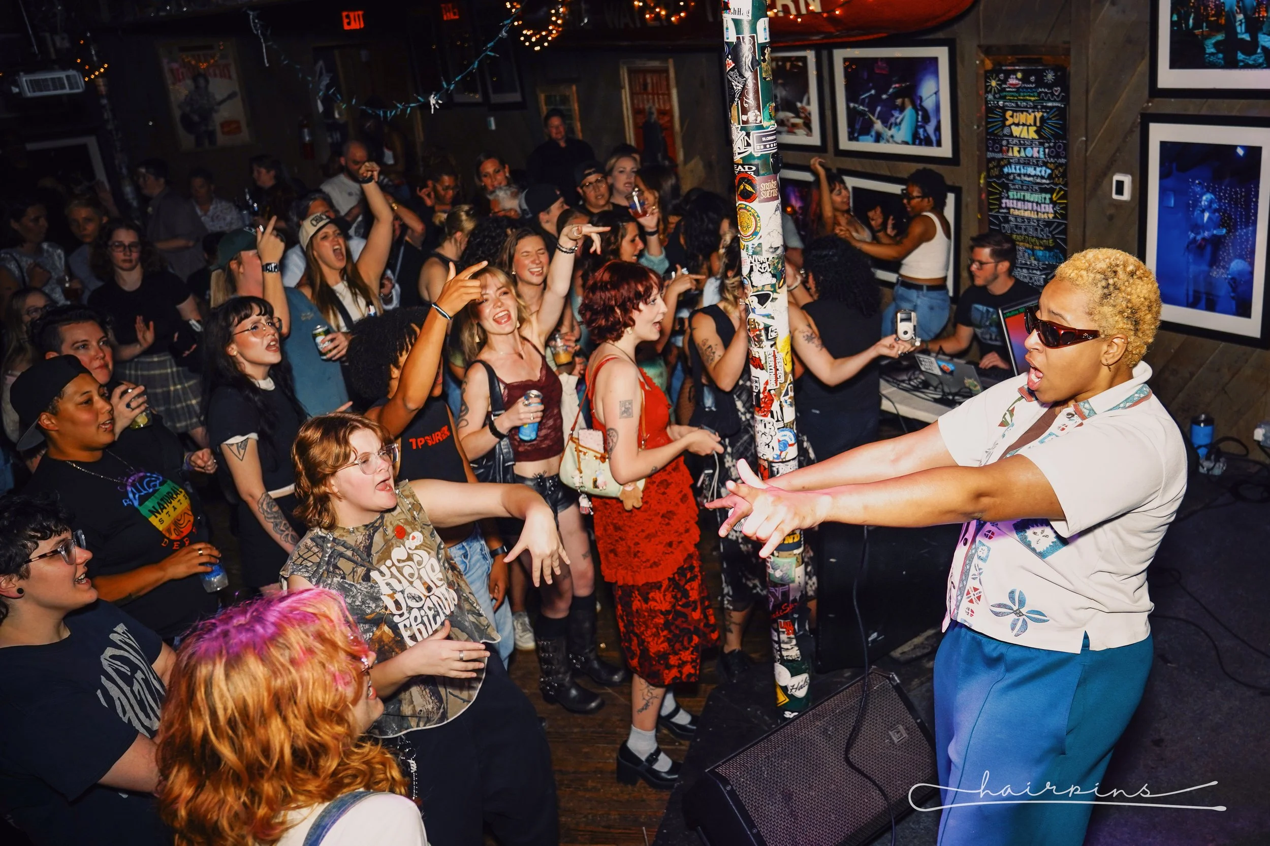 A lively concert with a singer performing on stage and an enthusiastic crowd of diverse young women dancing and singing along, inside a dimly lit venue decorated with photos and neon lights.