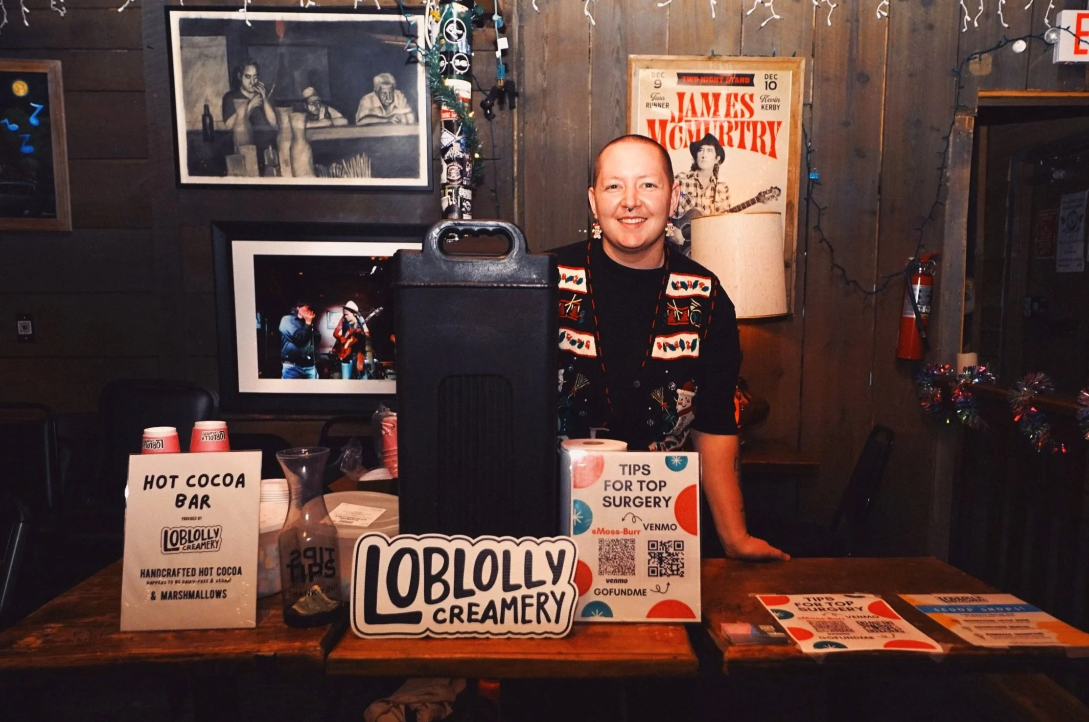 A smiling person standing behind a table at a booth for Lobolly Creamery in a cozy, decorated venue. The table has signs promoting hot cocoa and tips for top surgery. The background features wood-paneled walls with framed photos and posters, string lights, and snowflake decorations.