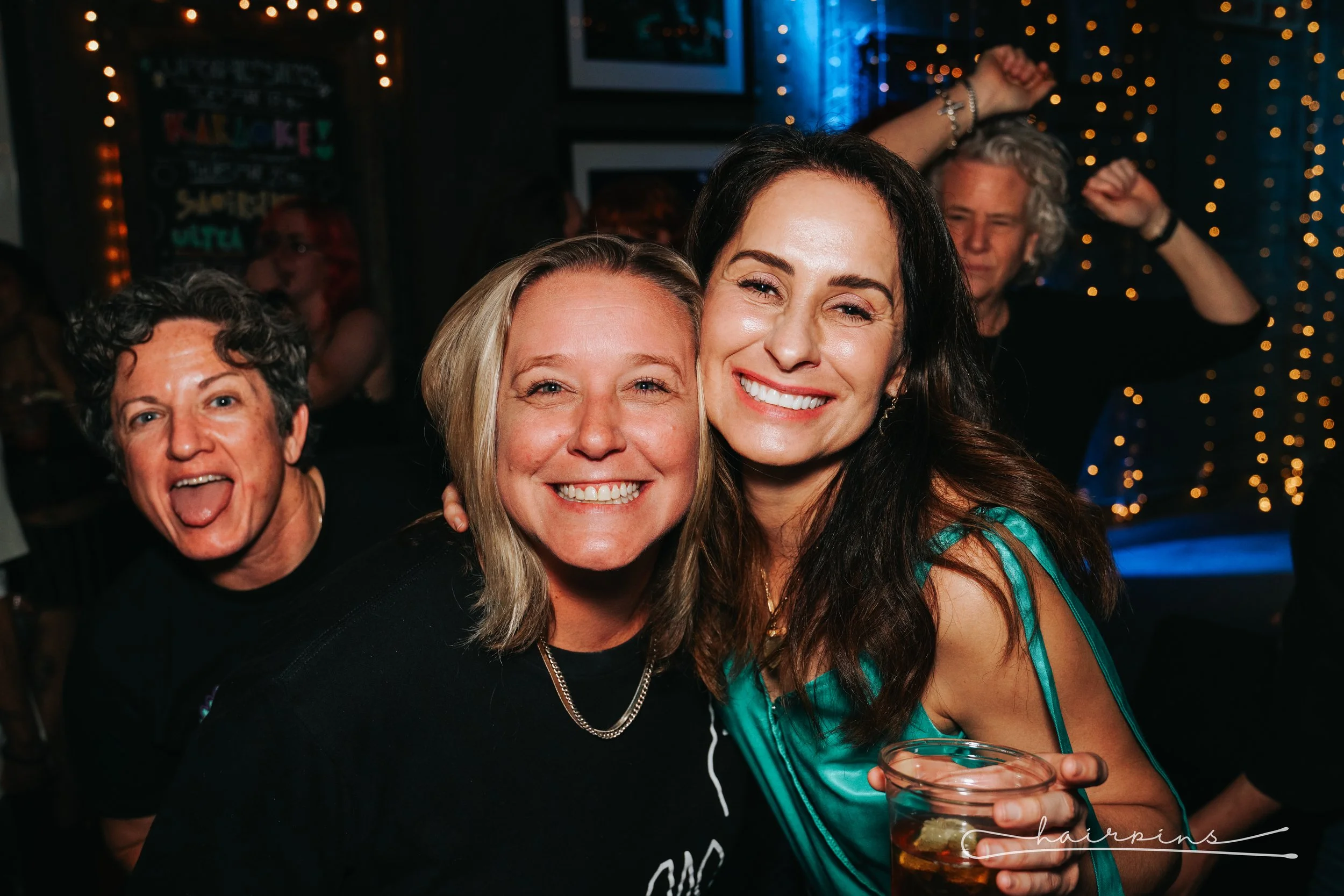 Group of people celebrating at a party, smiling and having fun in a dimly lit venue with string lights in the background.