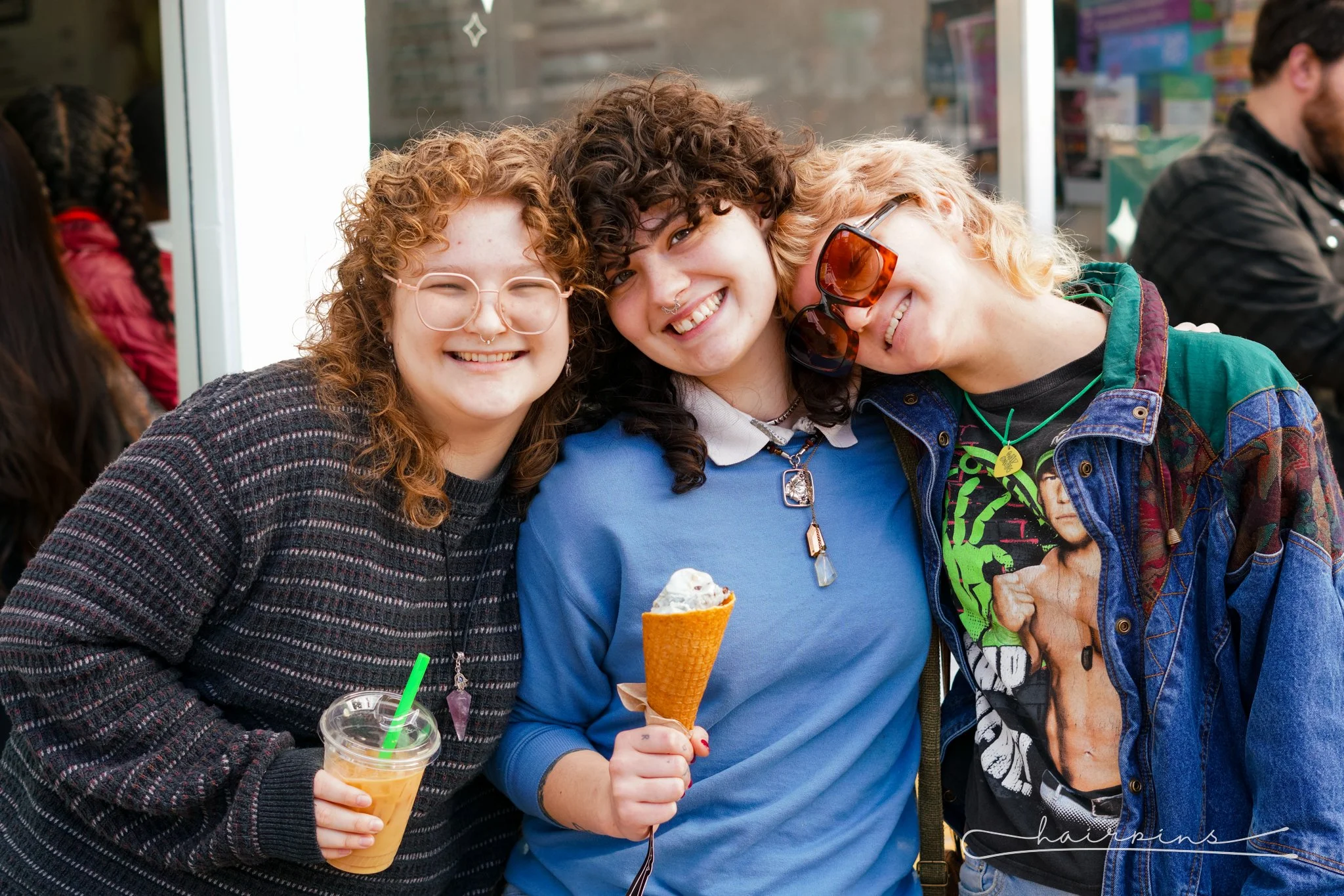 Three young women smiling and hugging, with one holding an ice cream cone, outdoors in an urban area.