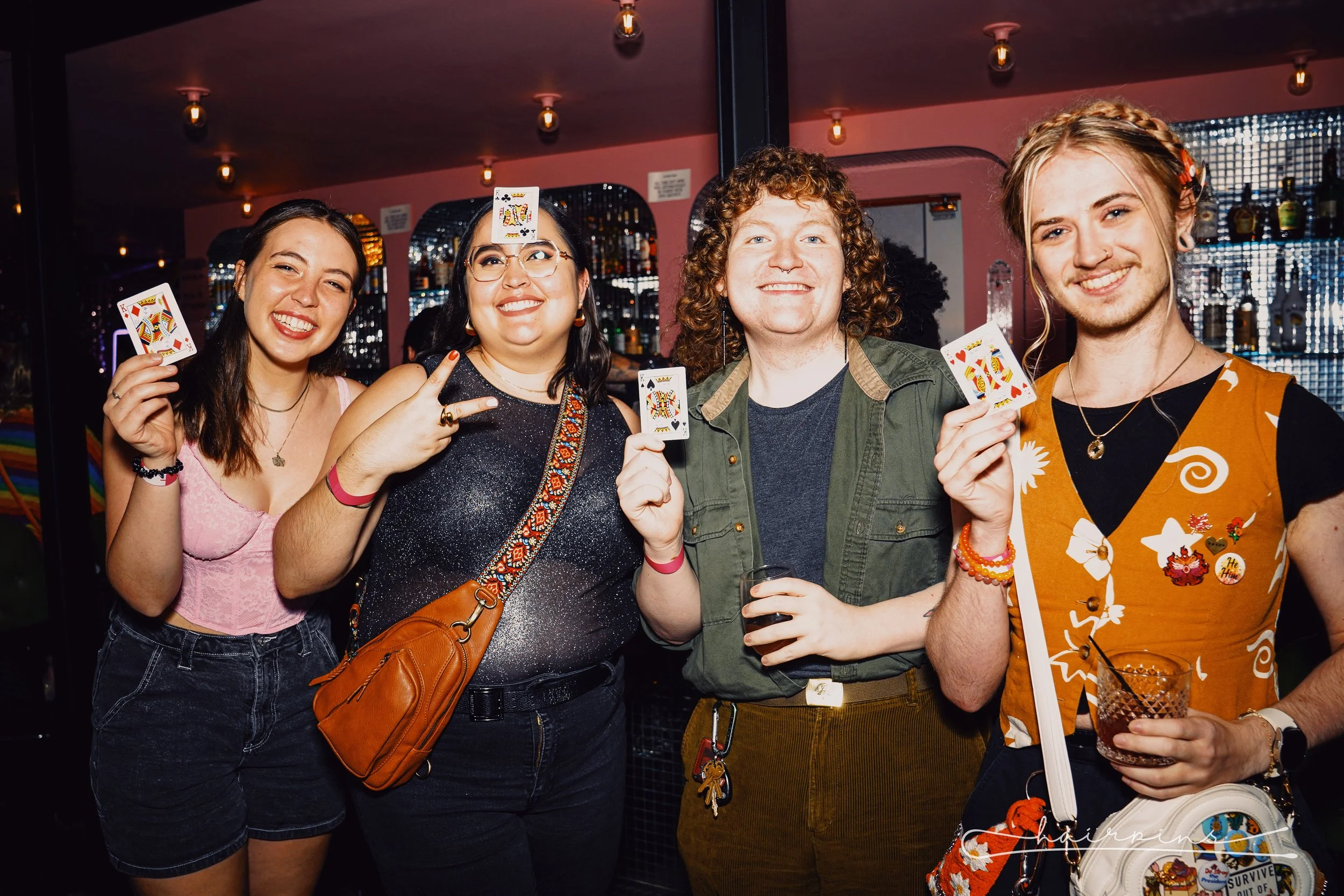 Group of four friends at a bar, smiling and holding playing cards, celebrating together.