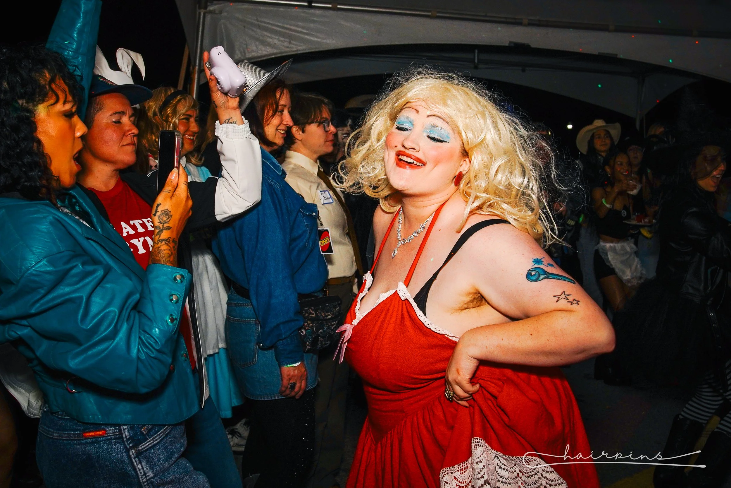 A woman with blonde curly hair dressed in a red dress, smiling and dancing at a party surrounded by people. She has tattoos on her arm and is wearing makeup with blue eye shadow.
