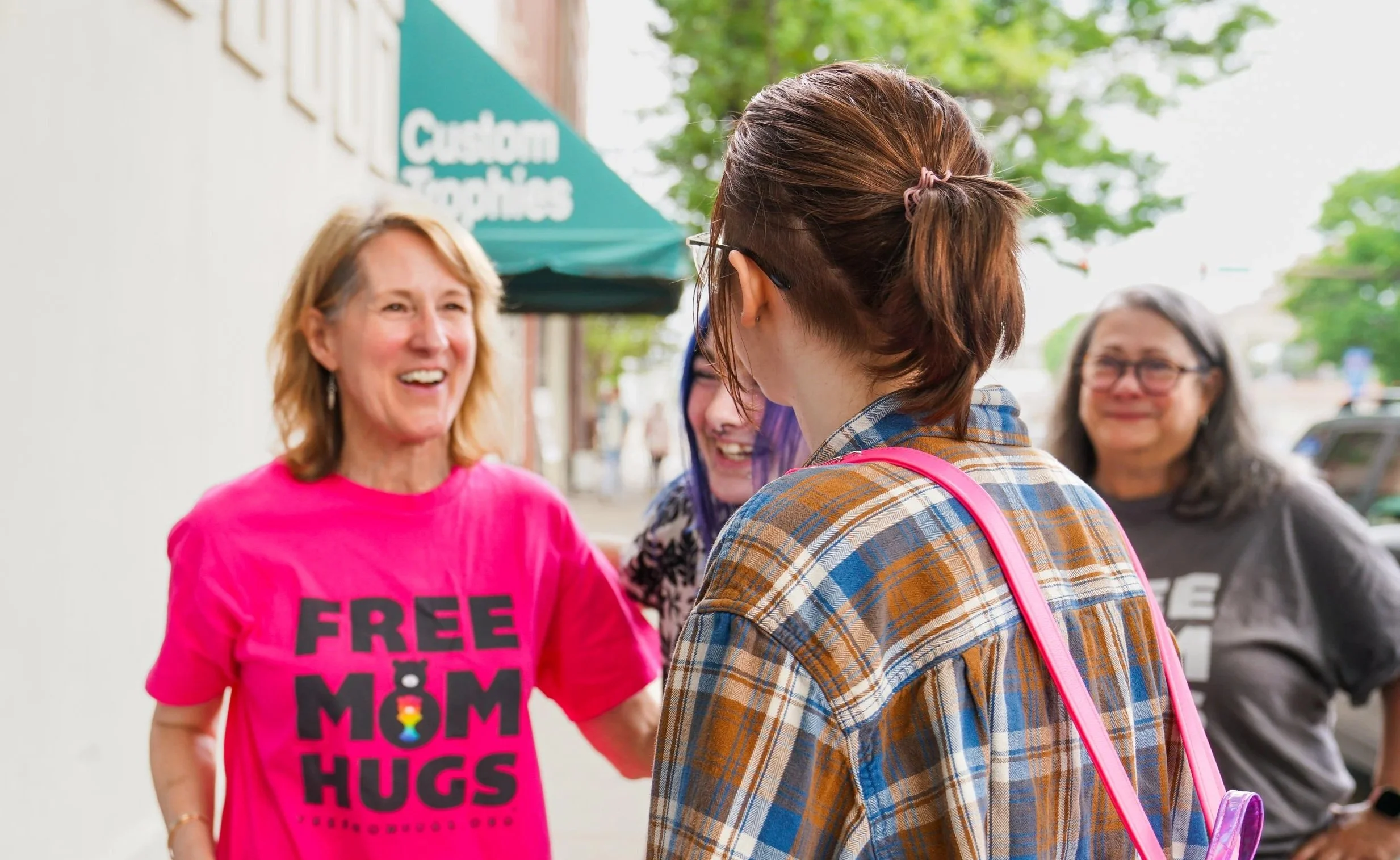 A woman wearing a pink t-shirt that says 'FREE MOM HUGS' greeting a group of women outside a store with a sign that says 'Custom Signatures' in the background.