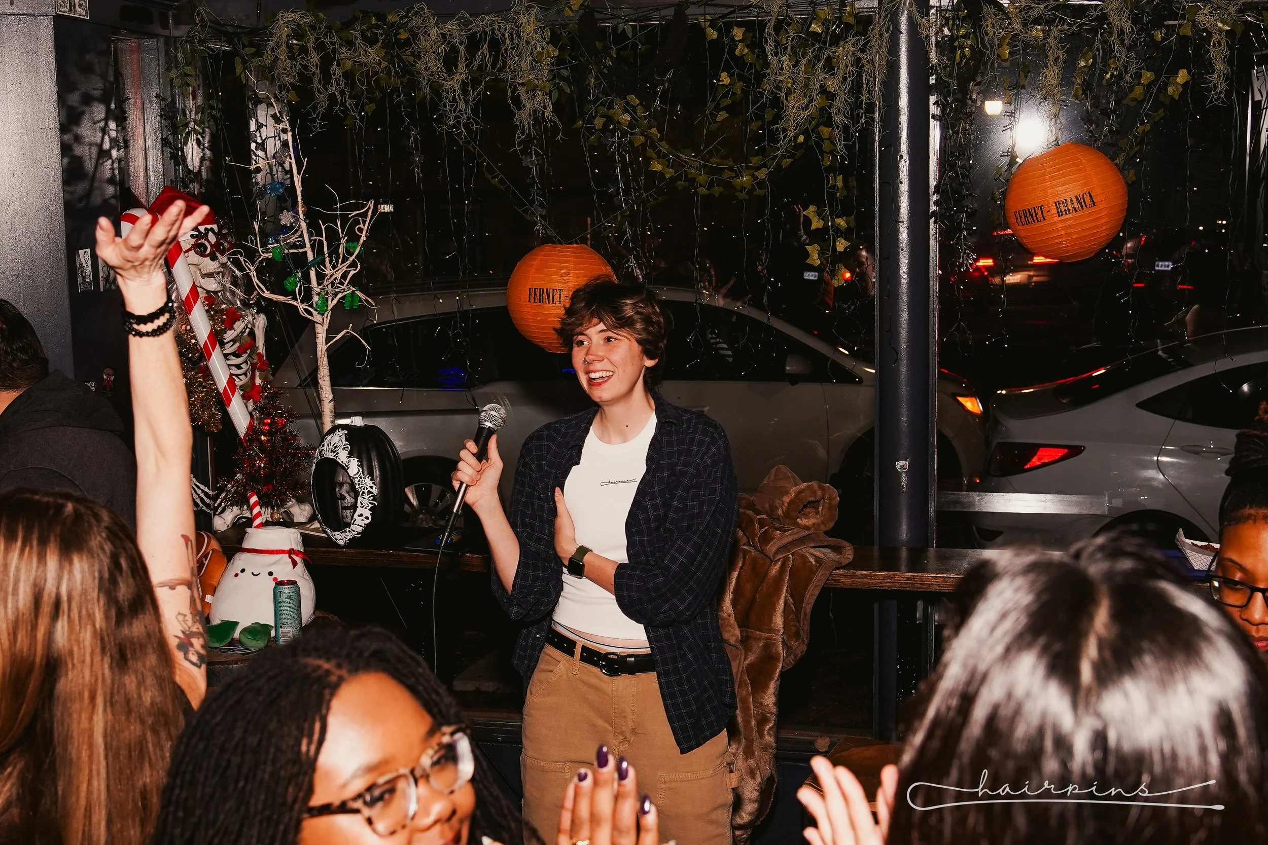 A woman with short hair and a checkered shirt holding a microphone and speaking at a social gathering inside a decorated space with Christmas and holiday decorations, including a small tree and orange lanterns that say 'Fernet-Branca', with an audience of diverse people in the foreground.