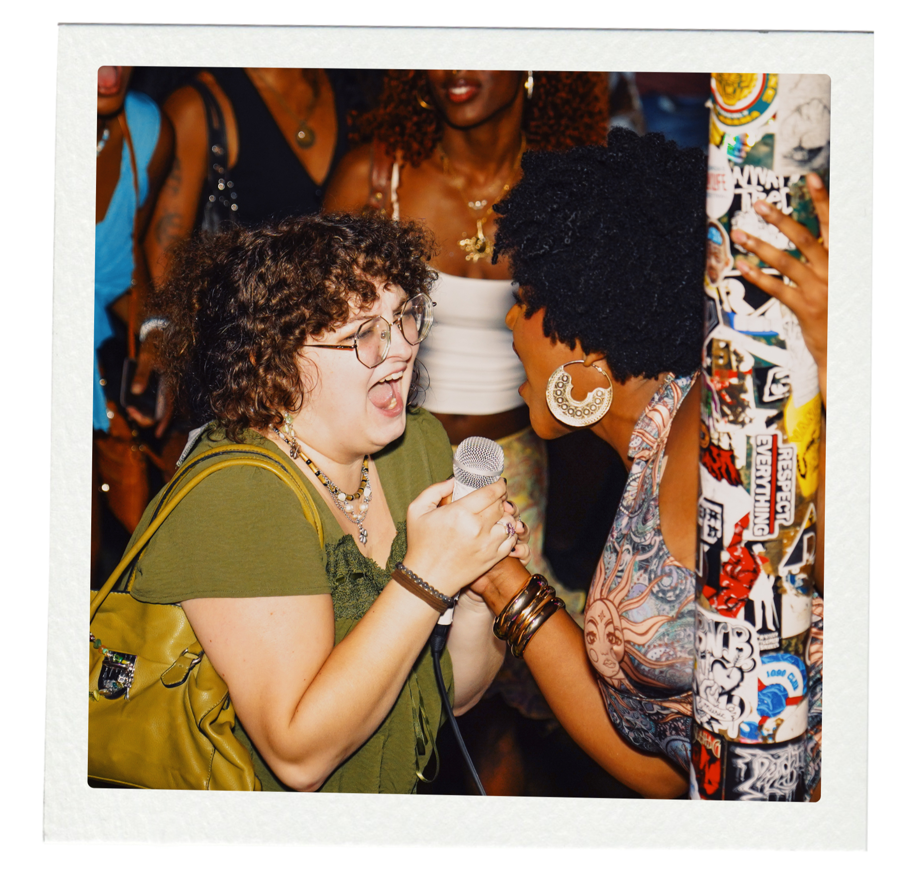 Two women are singing into a microphone together in a crowded setting, with one woman wearing glasses and a green top, and the other wearing large earrings and a colorful patterned shirt.