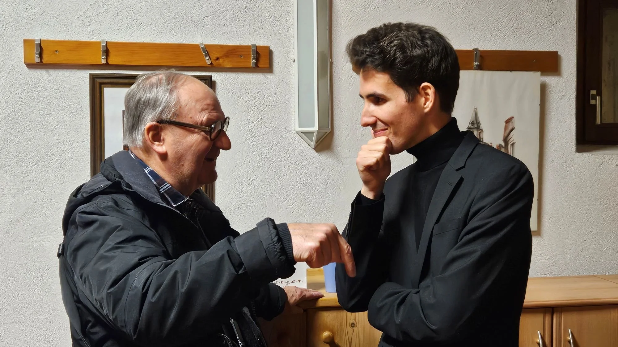 Ein älterer Mann und ein junger Mann unterhalten sich in einem Raum mit Holzdekoration an der Wand. Der ältere Mann zeigt auf den jungen Mann, der nachdenklich lächelt.
