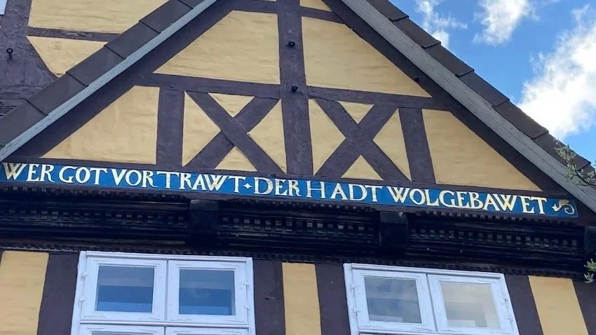 Ein Fachwerkhaus mit gelben und braunen Holzbalken, weiße Fenster und einem dekorativen Spruch in deutscher Sprache.