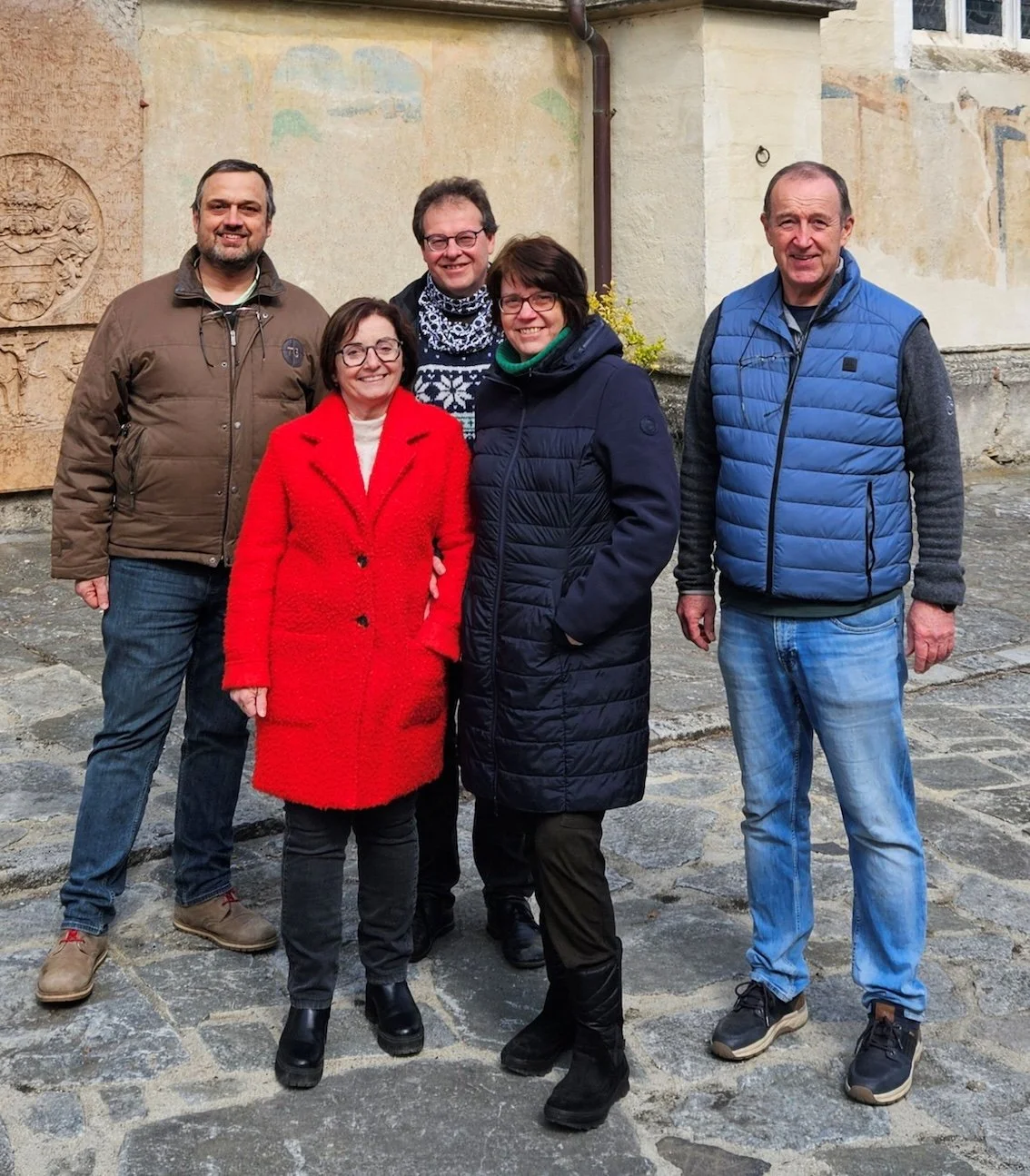 Das Team der Singerey. Fünf Menschen stehen auf einer gepflasterten Straße vor einer alten Gebäudewand, sie lächeln und posieren für die Kamera.