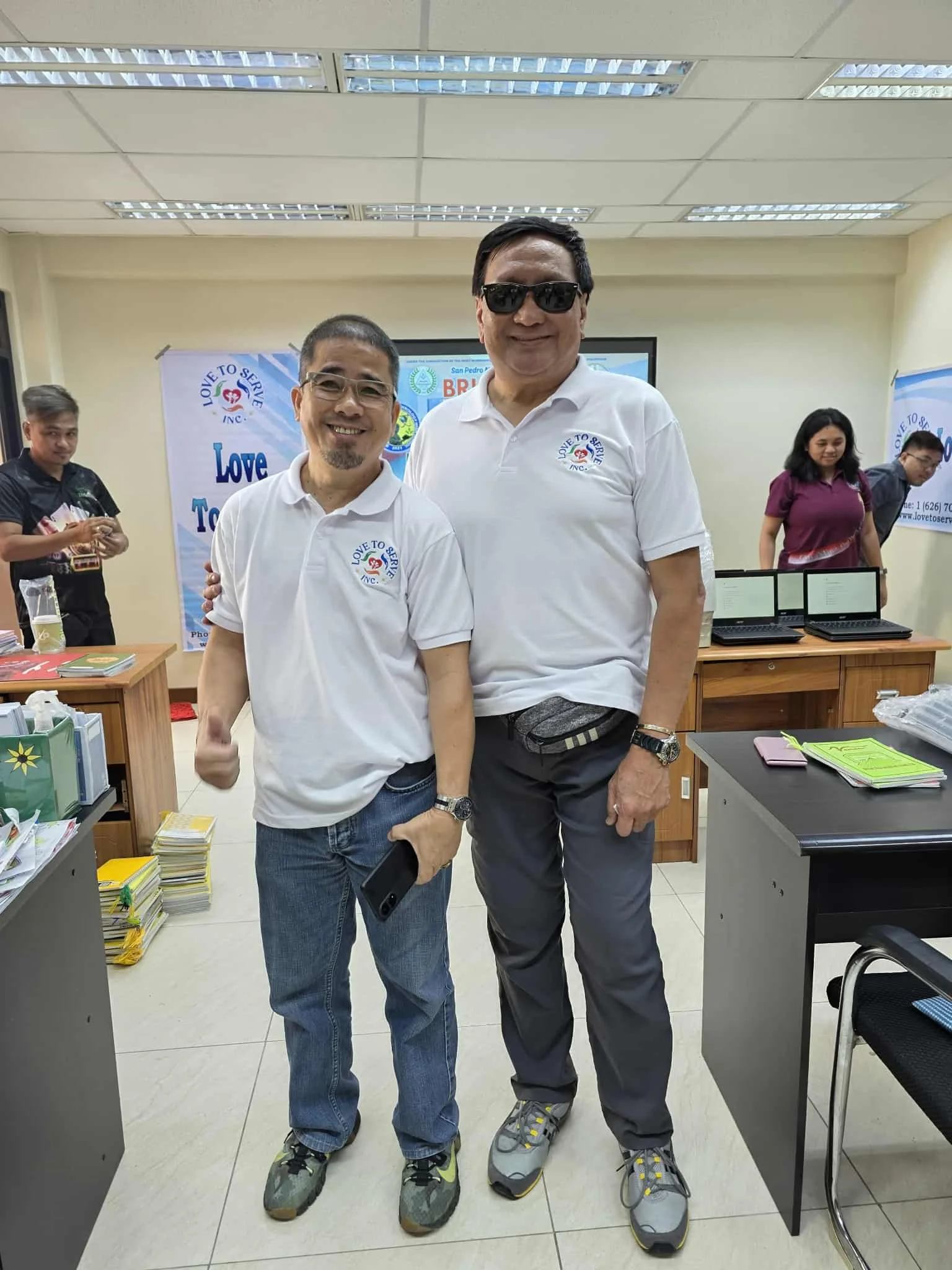 Two men wearing white shirts with a 'Love to Serve' logo stand together in an indoor office or meeting room, smiling at the camera. Multiple laptops, documents, and other people are visible in the background.