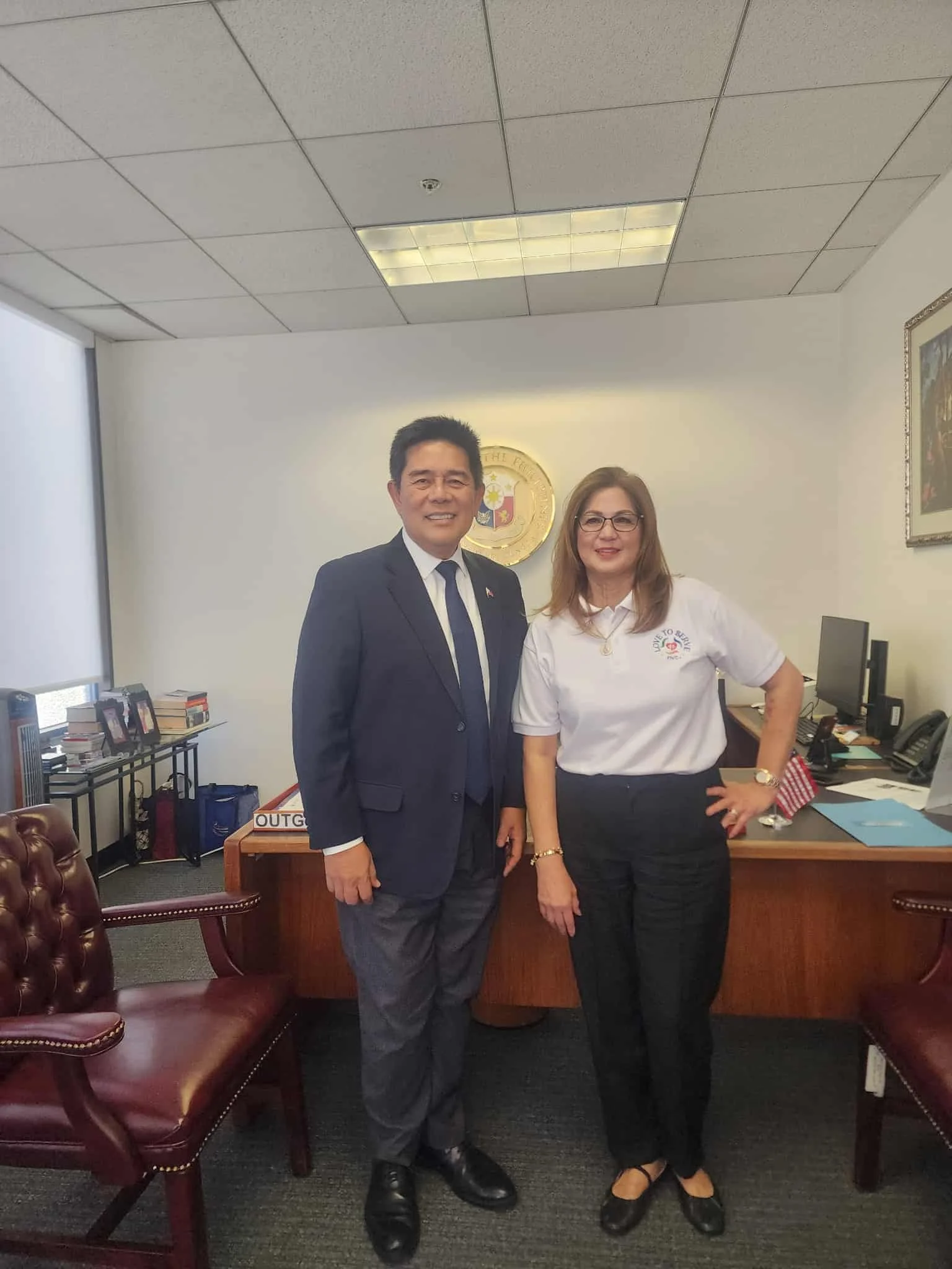 Two people standing in an office, one man in a suit and one woman in a white polo shirt and black pants, with a government emblem on the wall behind them.