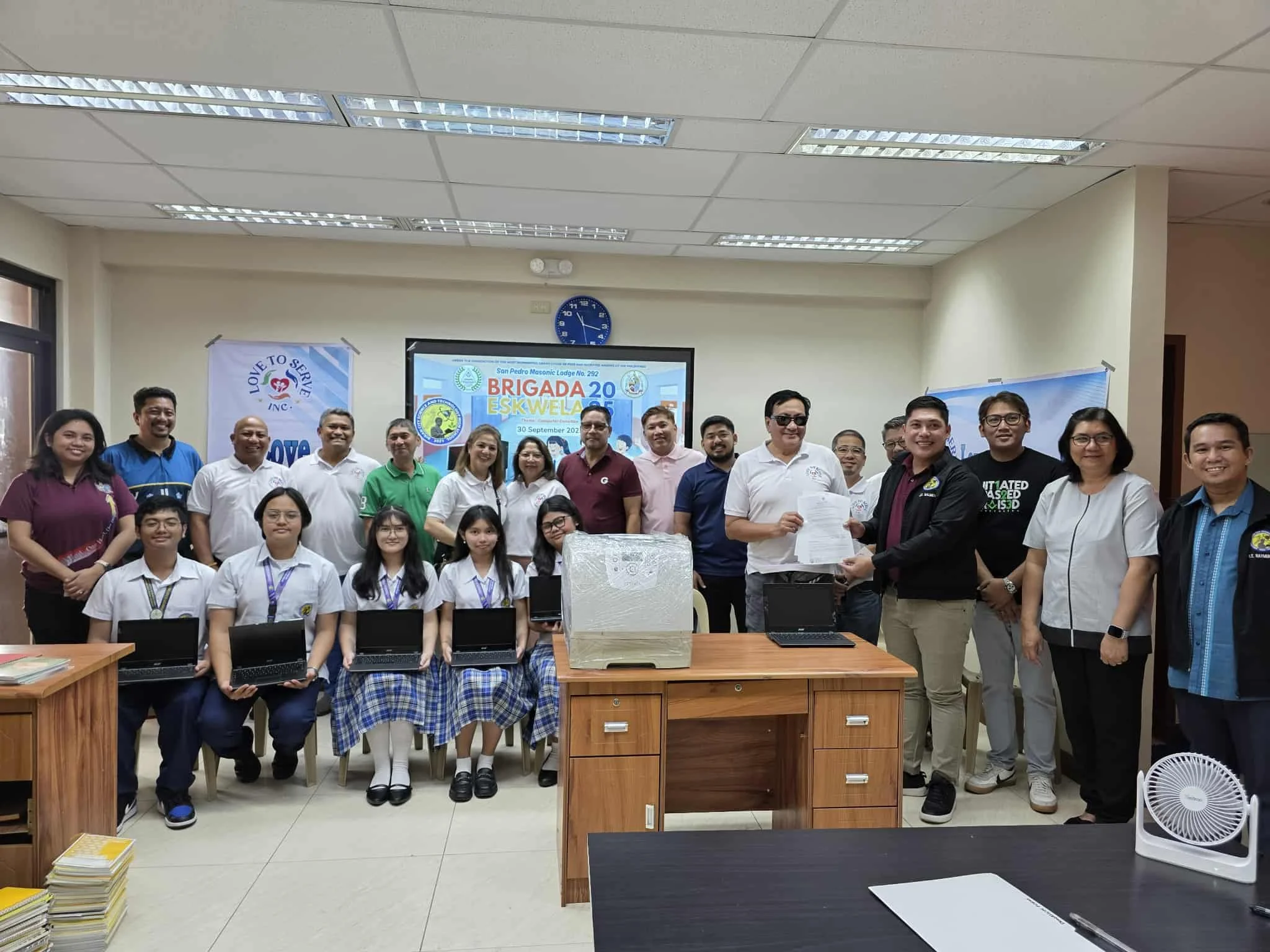 Group photo of students, teachers, and officials in a classroom during a ceremony for a school project. Some students hold laptops, and one man exchanges a document with another official. A presentation screen behind them displays the event details.