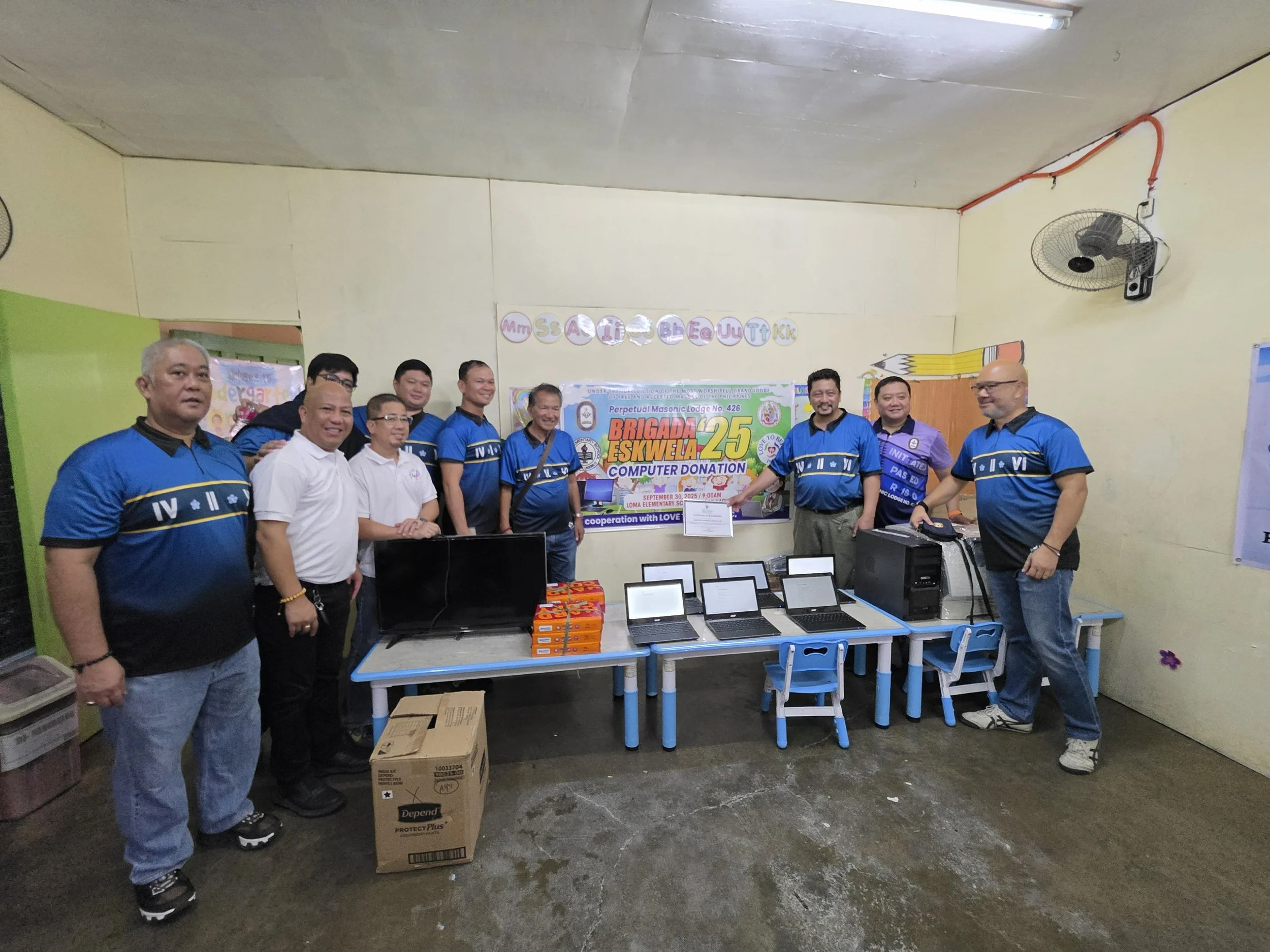 Group of men standing in a classroom with laptops and supplies for a computer donation event, with banners and colorful decorations on the wall.