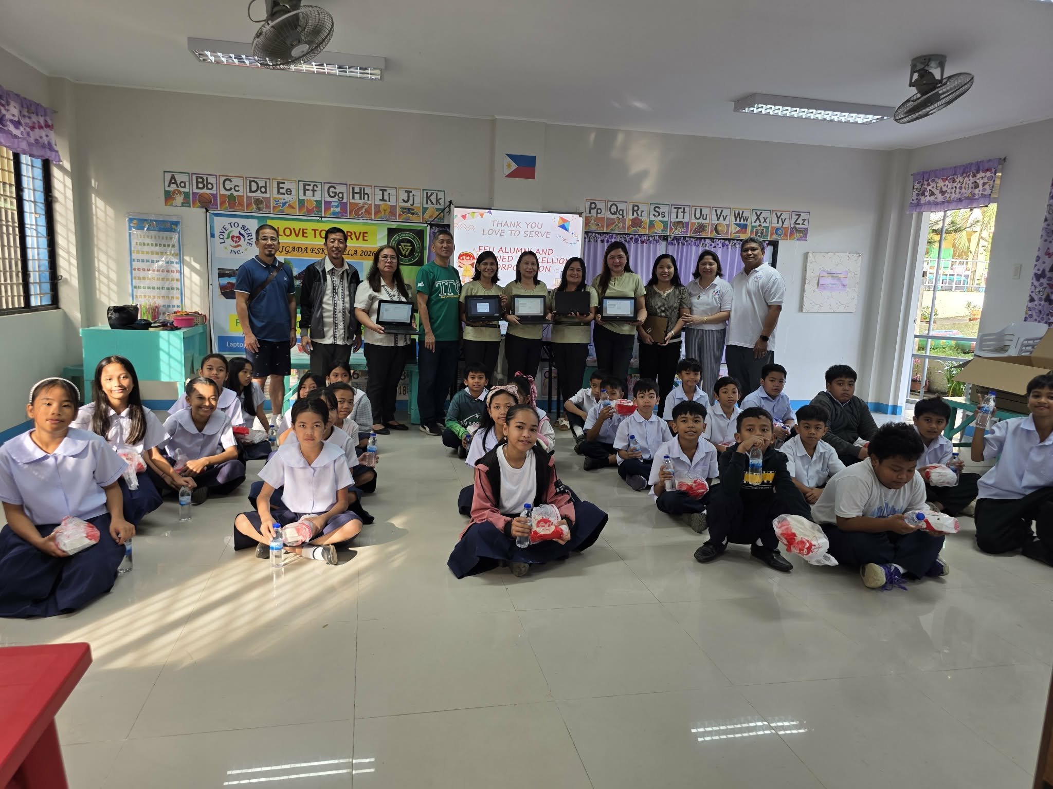 Classroom group photo with students sitting on the floor holding bottled drinks, and teachers standing behind them holding certificates or awards. The classroom has educational posters, alphabet charts, and a digital screen showing a thank you messag