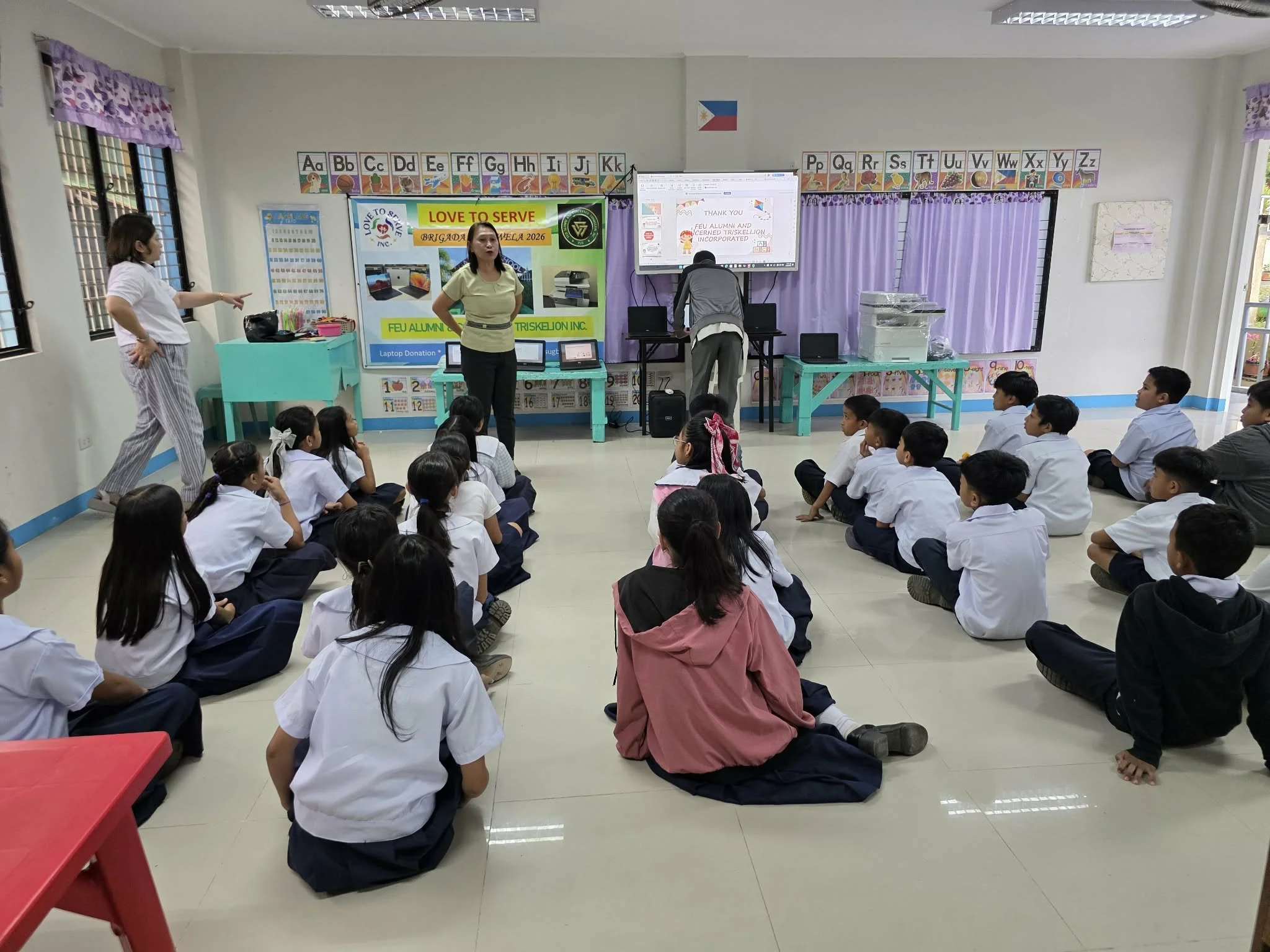 A classroom with students sitting on the floor, two teachers standing at the front, and a man setting up a projector. The classroom has colorful curtains, alphabet posters, and a Filipino flag.