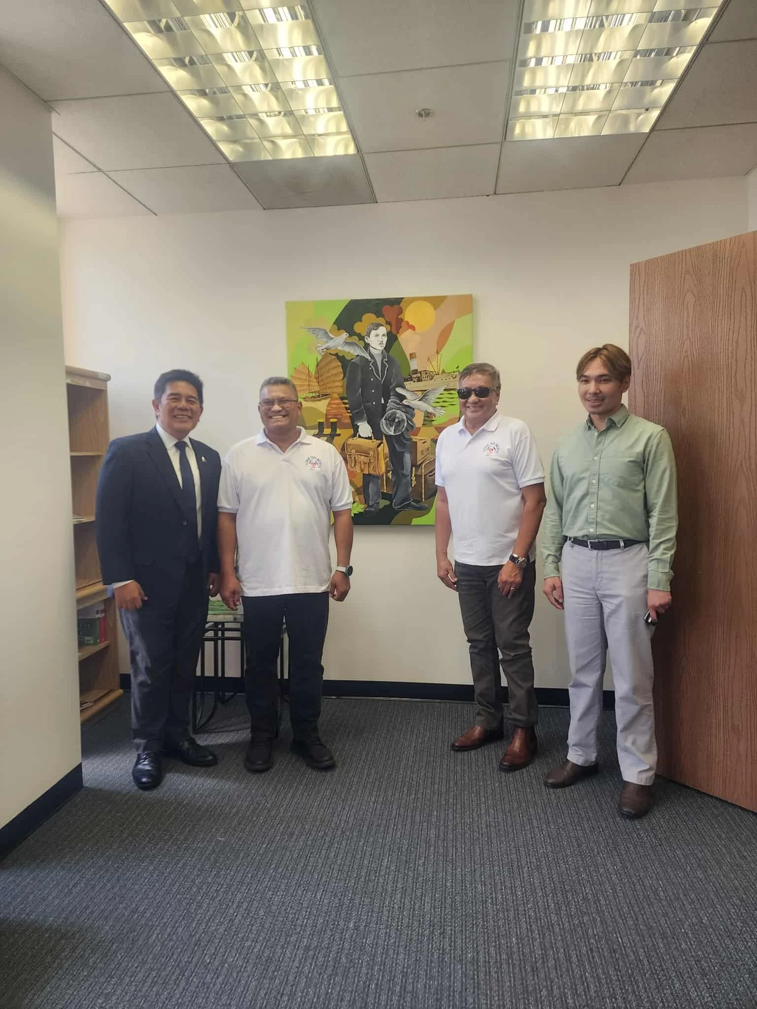 Four men standing in an office room, posing for the photo in front of a colorful painting of a woman holding a briefcase, with birds and a scenic background.