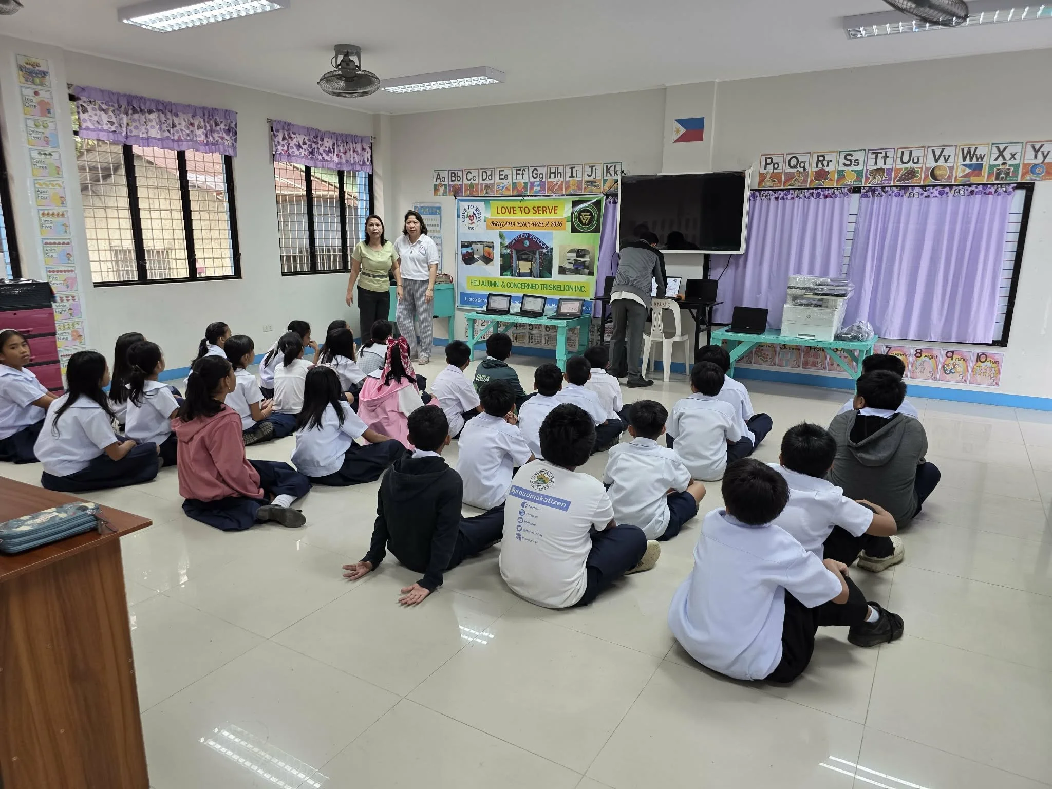 School classroom with students seated on the floor, facing a presentation area with teachers and a display board.