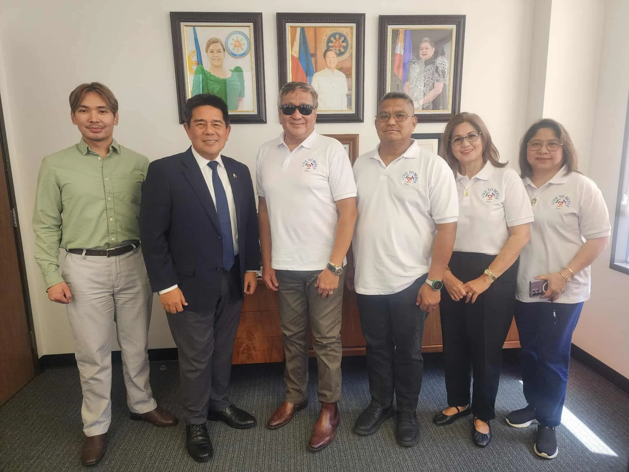 Seven people standing together inside a room, five men and two women, posing for a group photo. The background features framed portraits of three women on the wall.