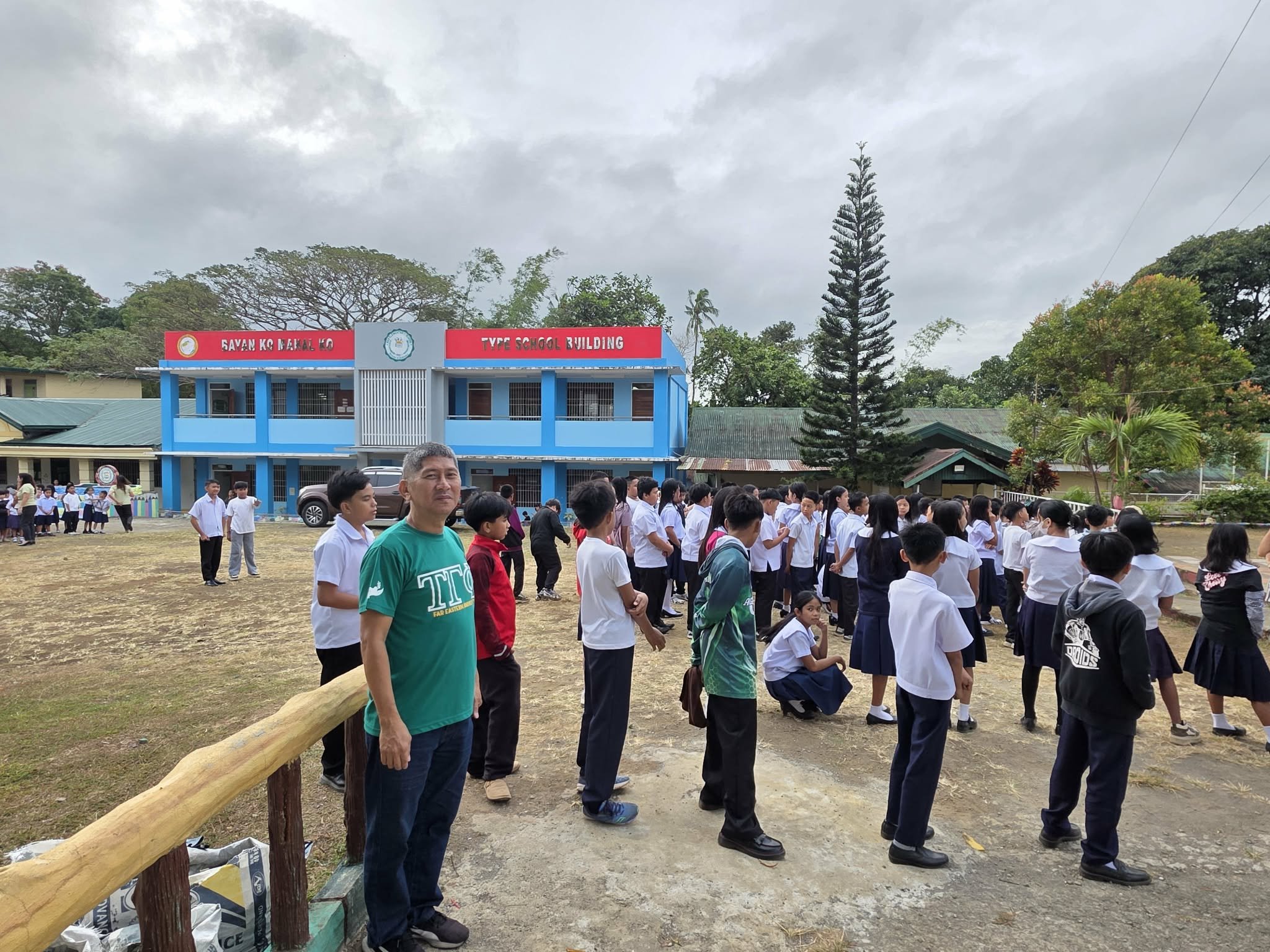 Students in school uniforms standing in line outside a blue school building with a red sign, on a cloudy day with trees in the background.