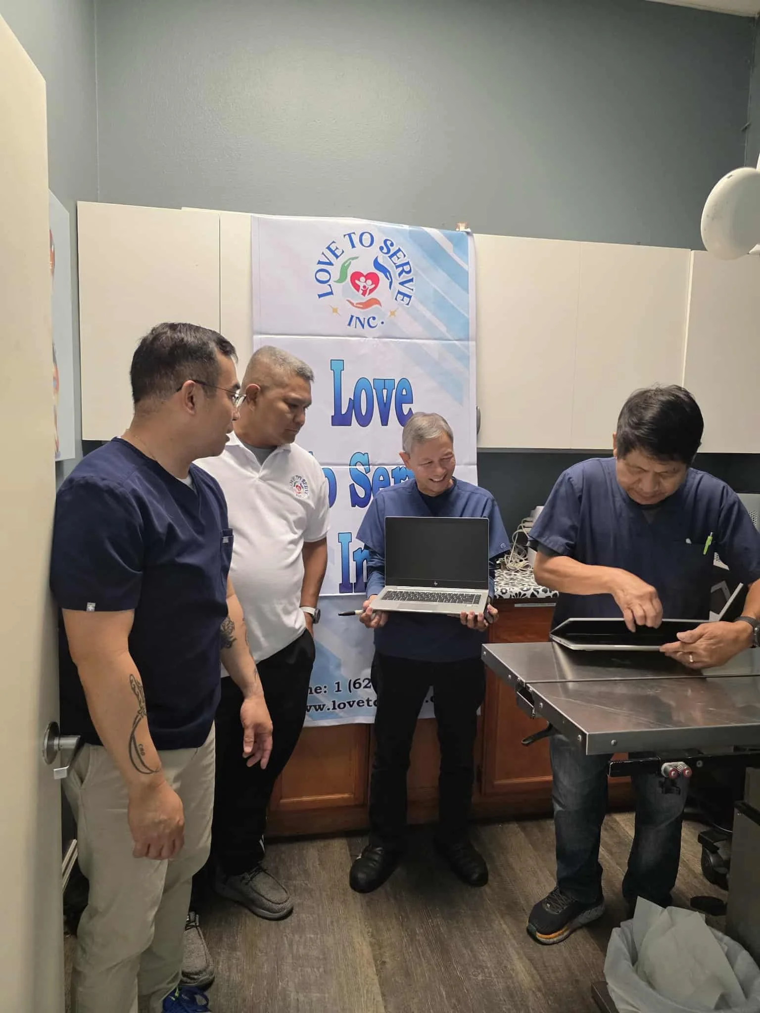 Four men standing in a room with a banner that reads 'Love to Serve' behind them, one holding a laptop, and another looking at a tablet.