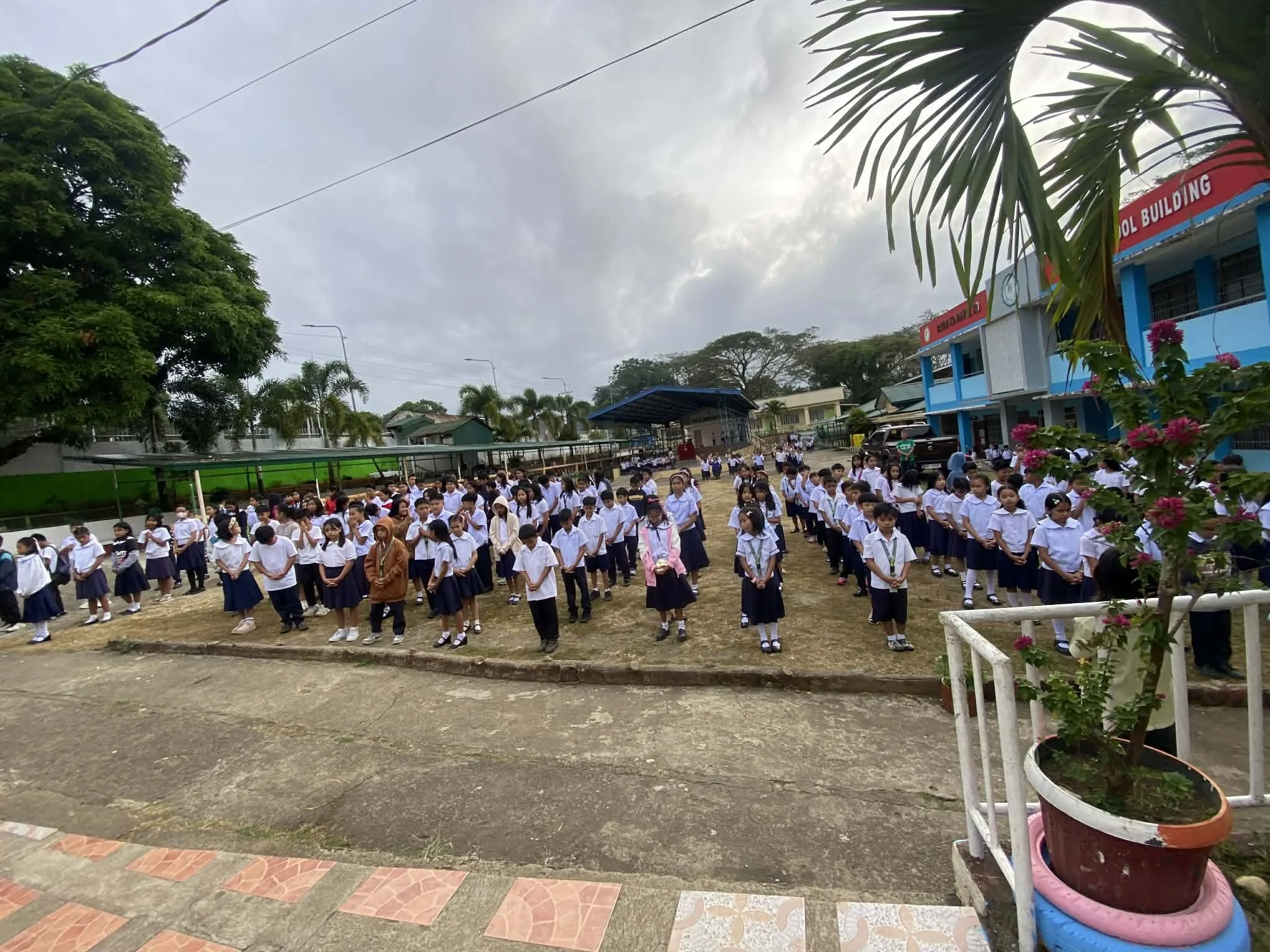 School children standing in lines outdoors on a cloudy day, dressed in uniforms, some wearing face masks, in front of a school building with a sign that reads 'SCHOOL BUILDING.'