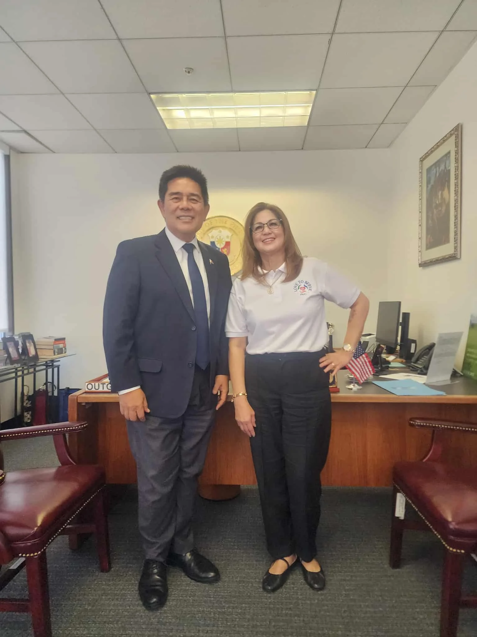 A man in a navy suit and a woman in a white shirt and black pants standing in an office, smiling at the camera.