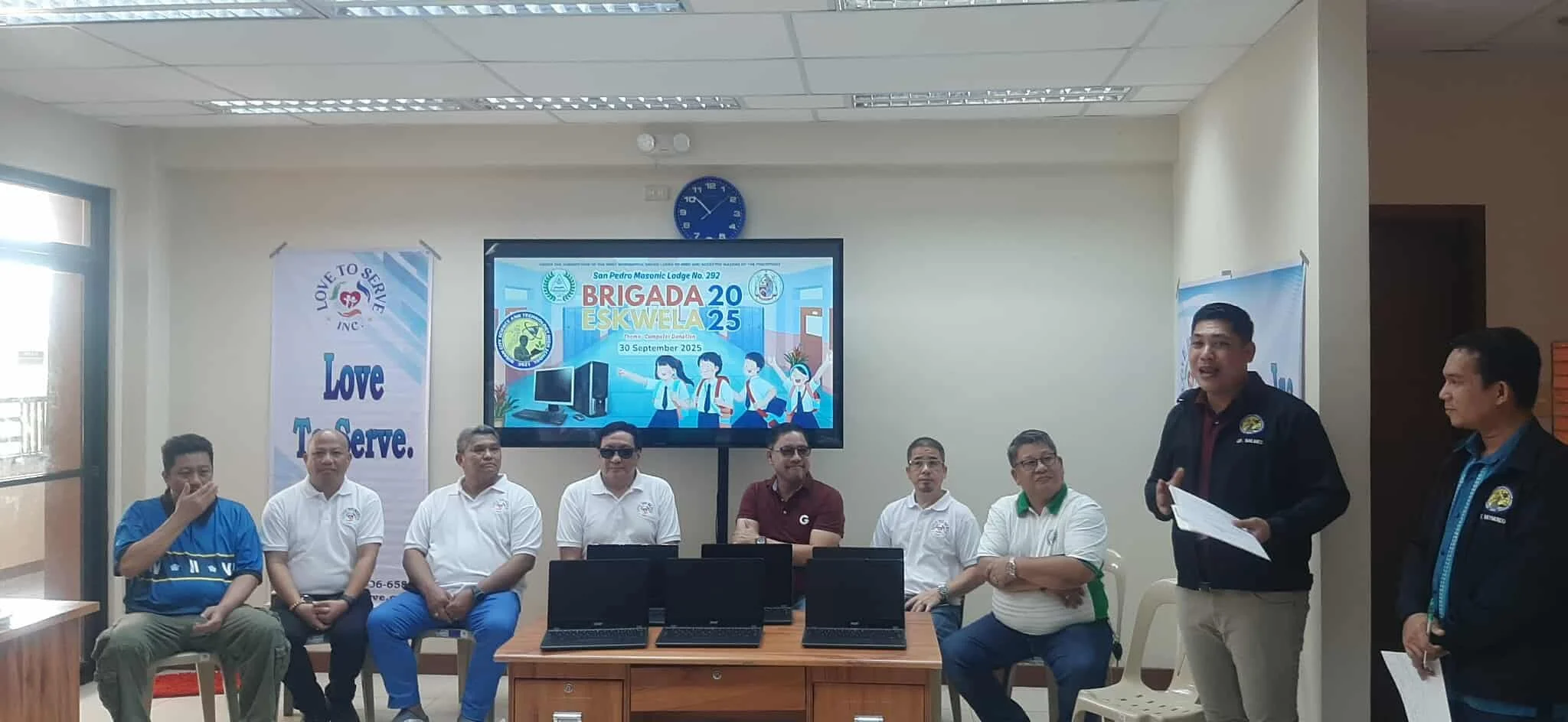 A group of men seated and standing in a conference room during a presentation or meeting, with a large screen behind them displaying a colorful banner for a school event on September 30, 2025, and a banner on the left with the text "Love To Serve."