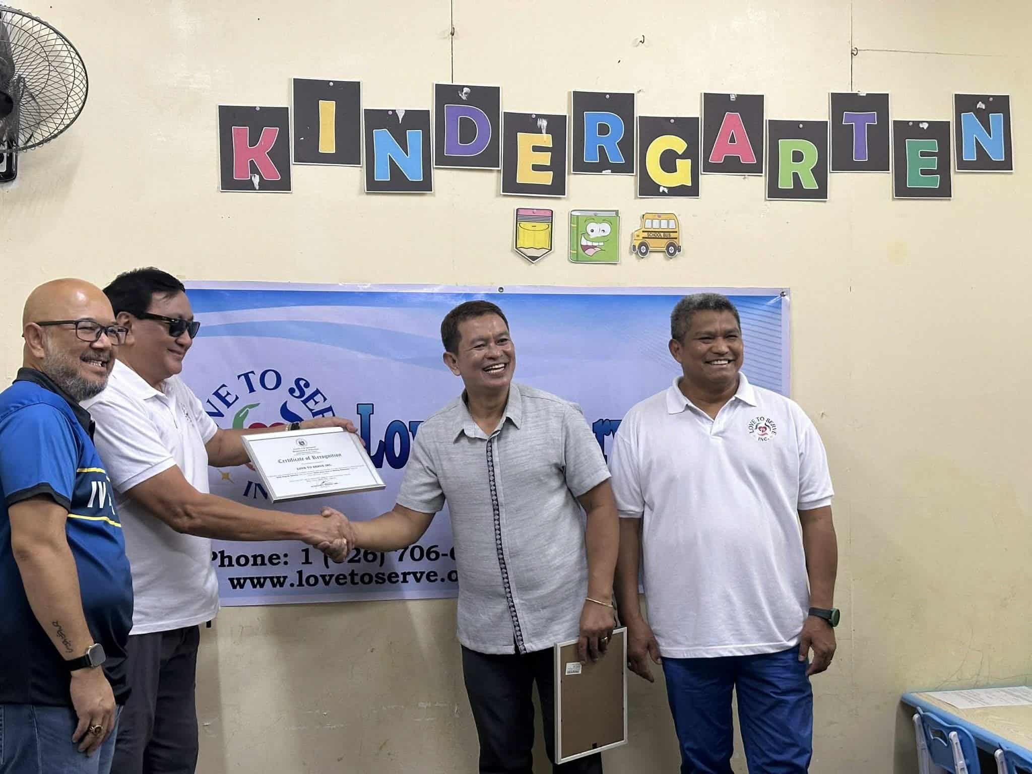 Four men standing in front of a blue banner that reads "Welcome to Serve". Two of them are shaking hands, one of whom is holding a certificate. The background has a colorful "Kindergarten" banner with letters and small illustrations.