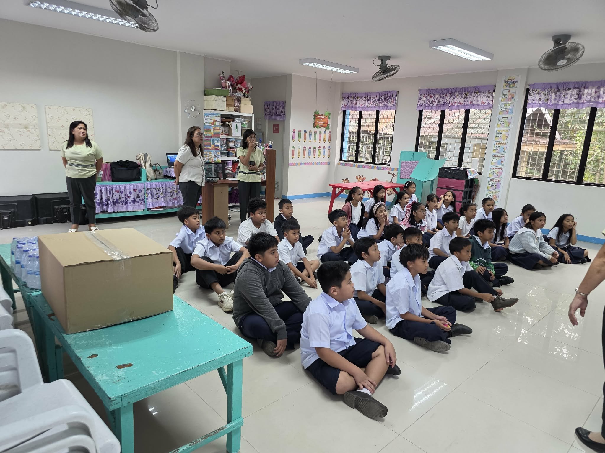 A classroom filled with young students sitting on the floor, facing forward. Behind them are teachers standing near a desk and shelves, with colorful decorations and windows in the background.