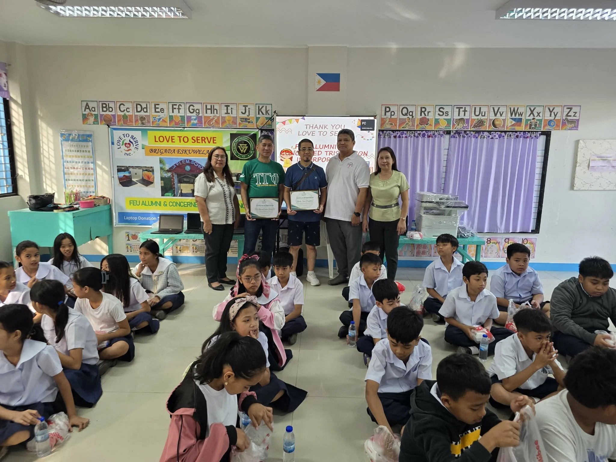 School classroom with students seated on the floor and teachers standing at the back holding certificates, celebrating an event related to alumni and community service, with Philippines flag and educational posters visible.
