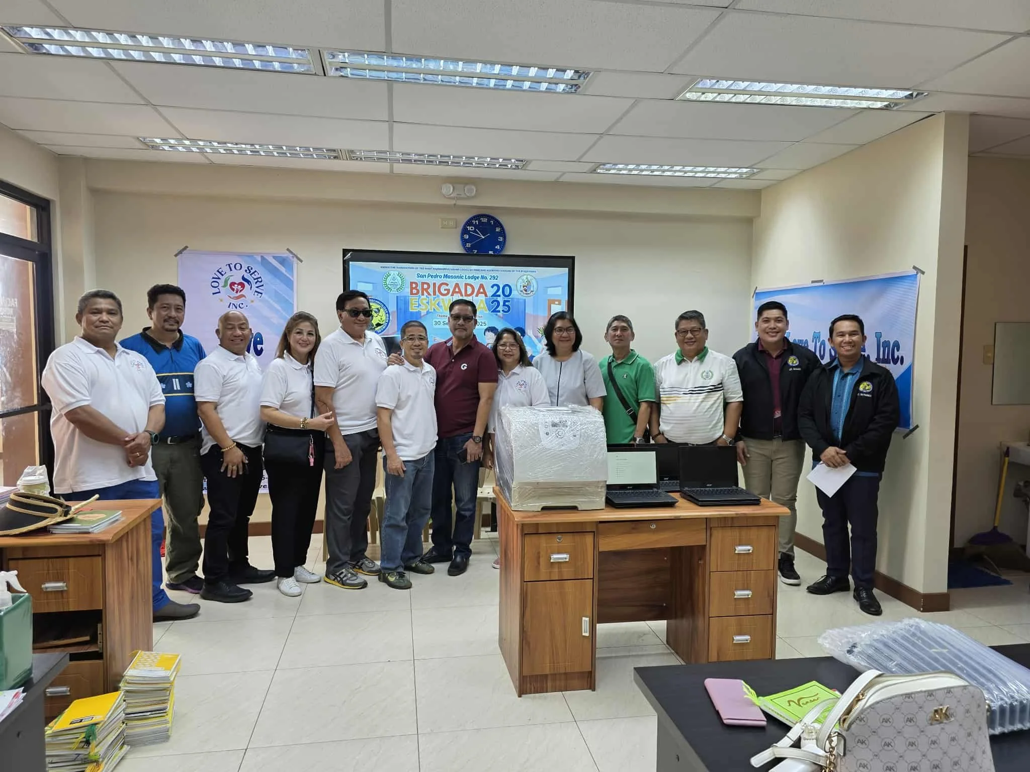 A group of 14 people standing in a room with a presentation screen behind them. The screen displays text related to a Brigada Eskwela event. The room has fluorescent lighting and windows on the left side. The group is smiling and dressed in casual to