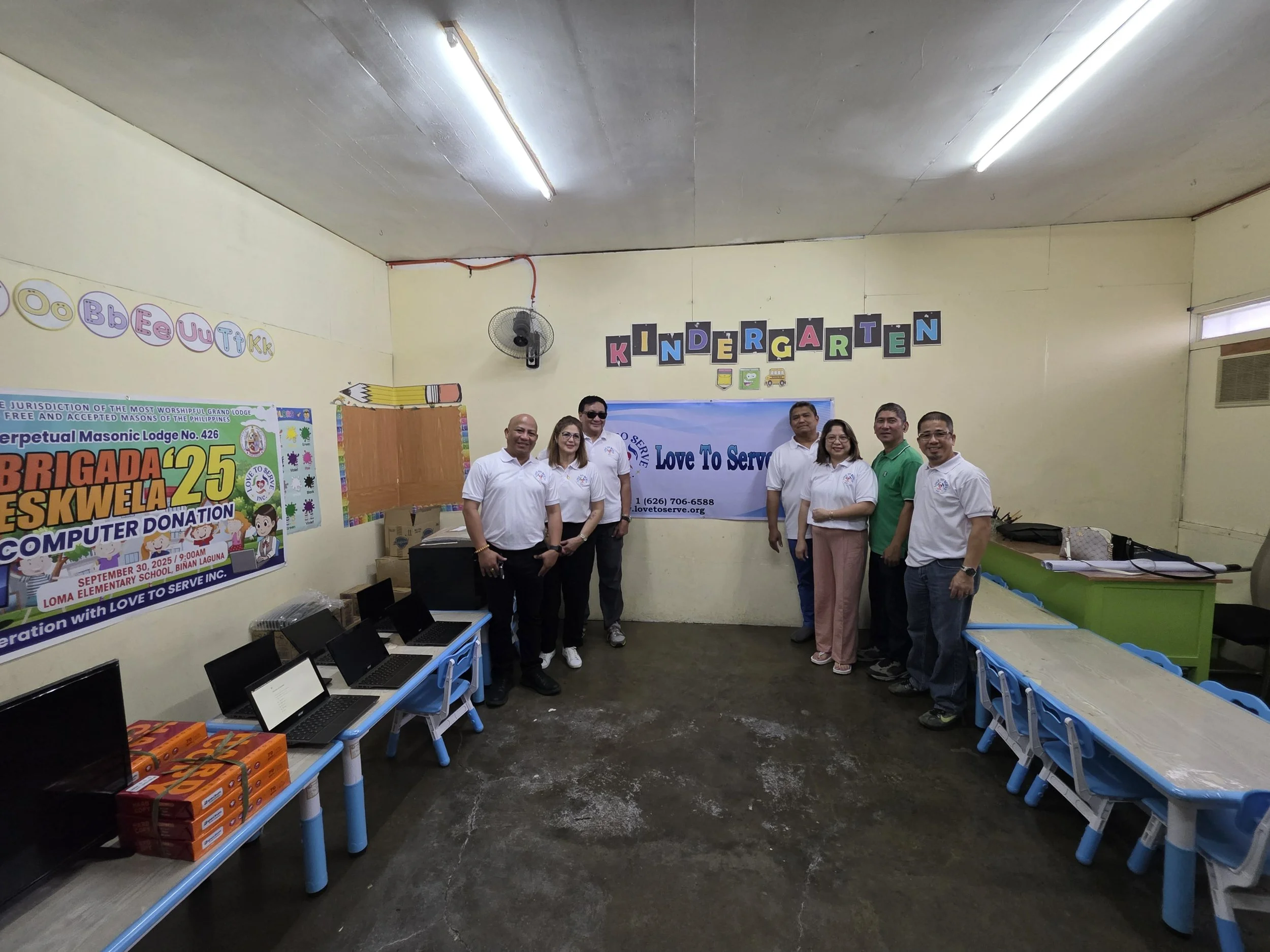Group of eight adults in a classroom with computers and a banner that reads 'Love To Serve.'