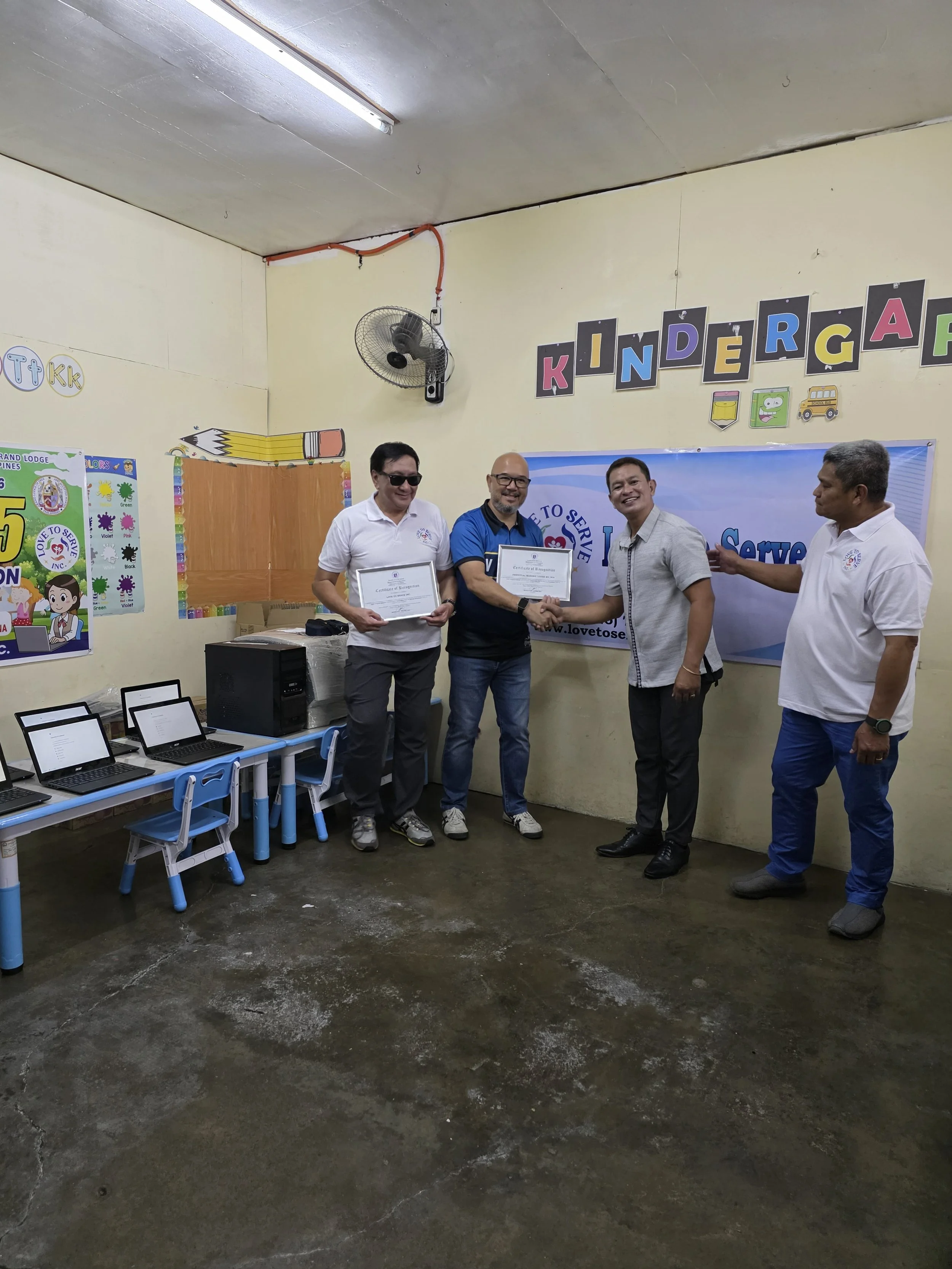 Four men in a classroom, with two holding certificates and shaking hands, in front of a "KINDERGARTEN" sign, celebrating an achievement or event.