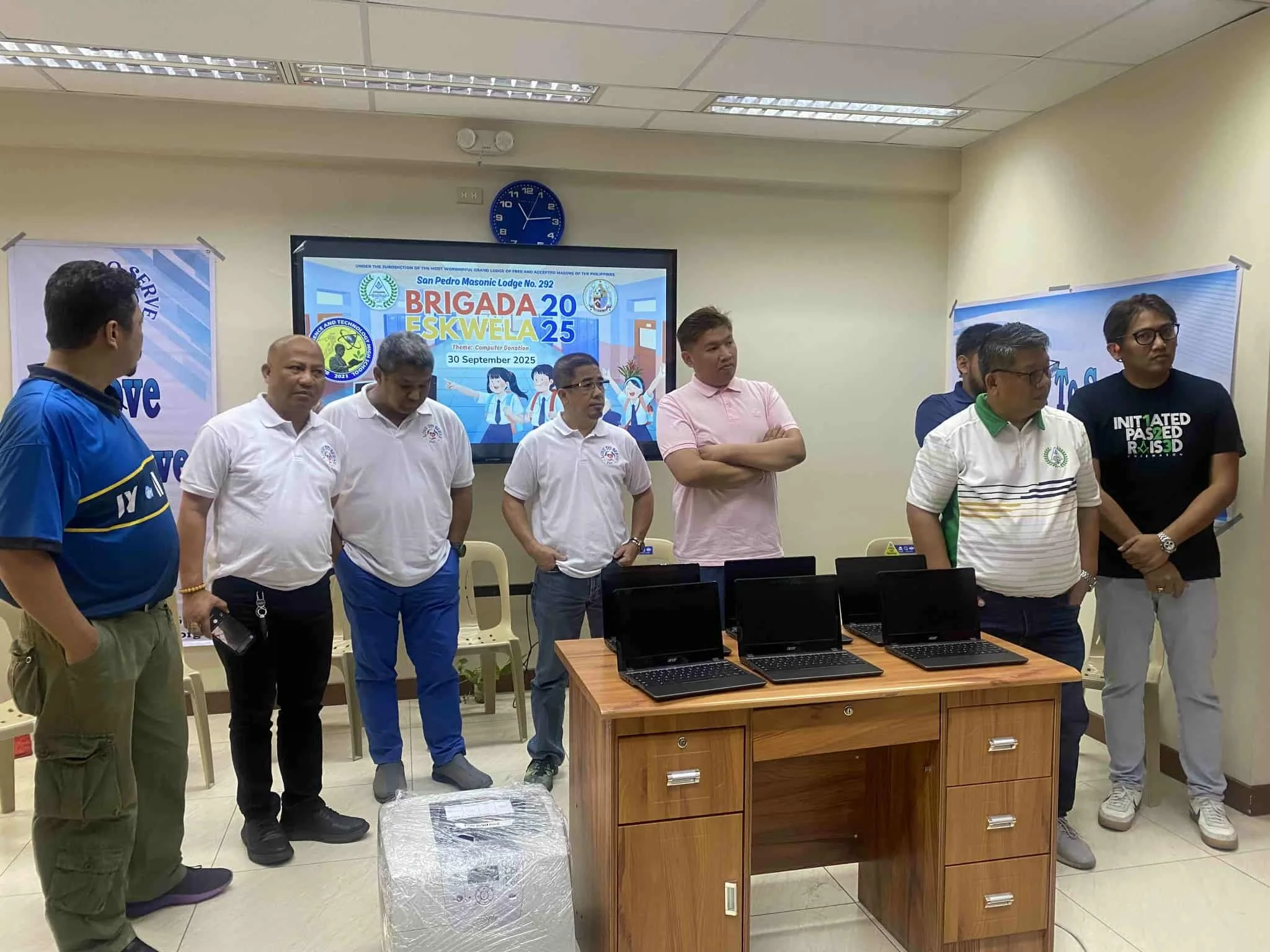 A group of seven men standing behind a wooden table with four laptops. They are in a room with a large digital screen and banners. The digital screen displays the text "Brigada Eskwela 2025" and includes logos, a date of September 30, 2025, and carto