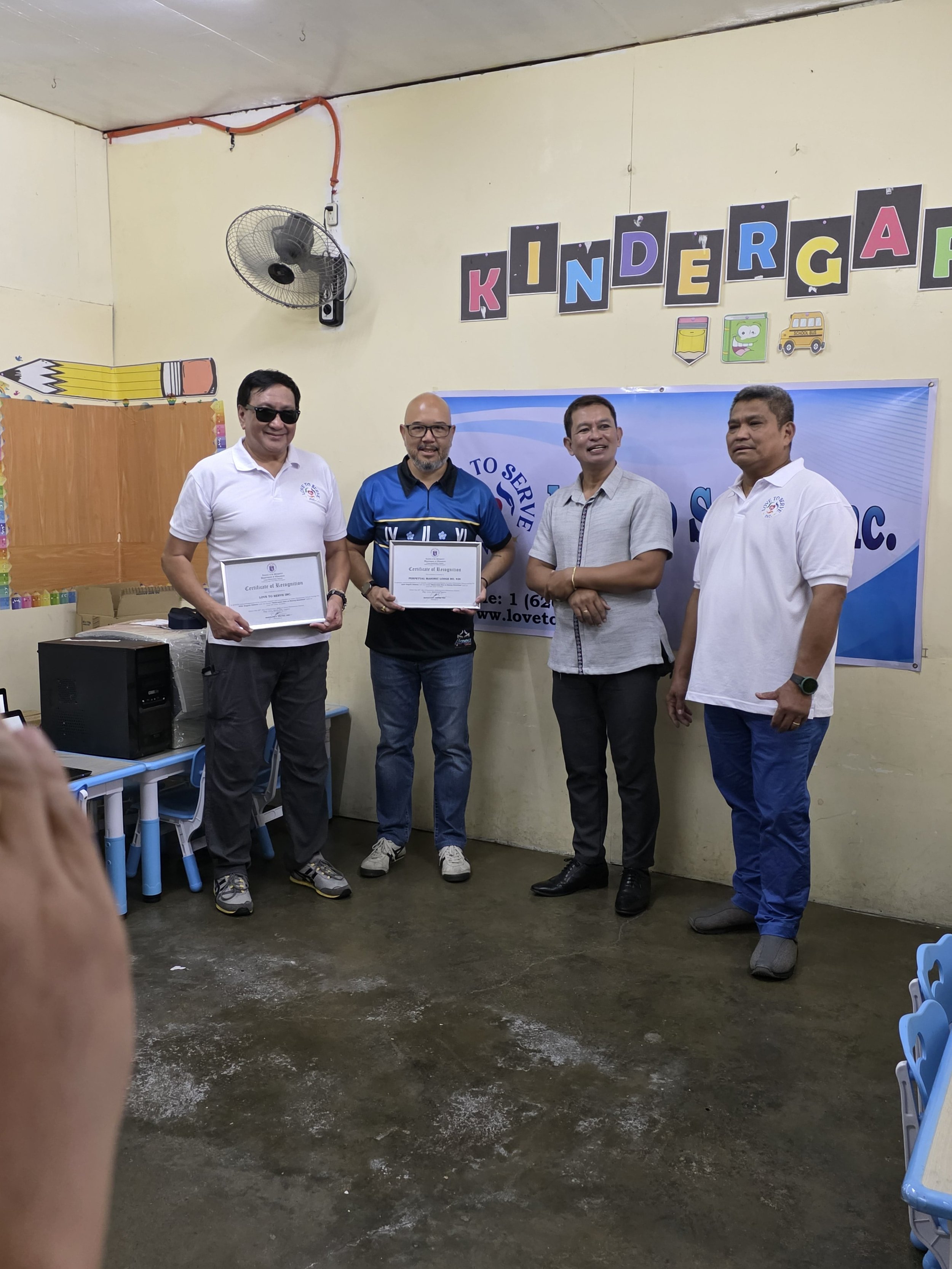 Four men standing in a classroom, two holding certificates, against a wall with a birthday decoration spelling 'KINDERGARTEN' and a banner, with chairs and a table nearby.
