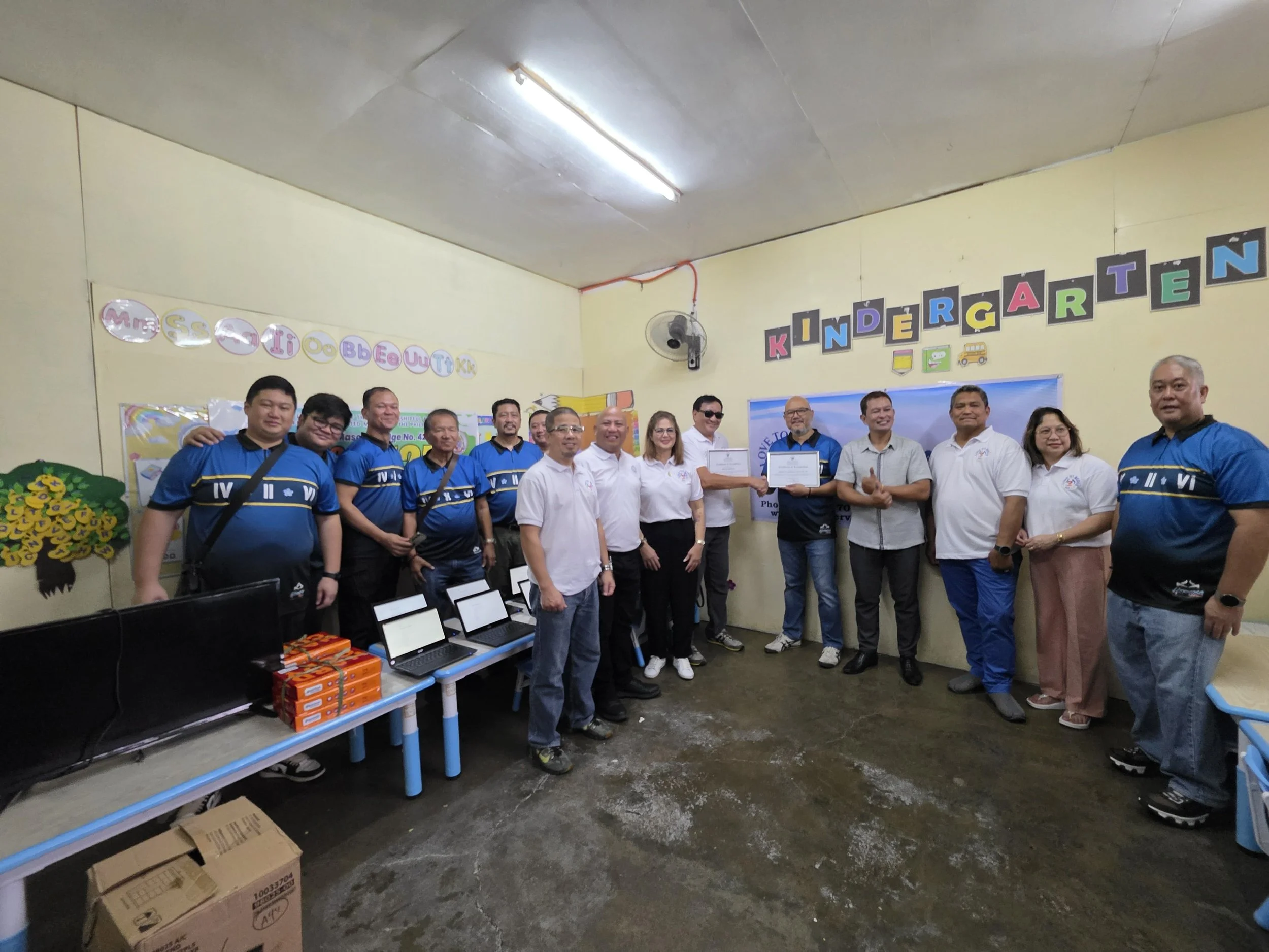 Group of people standing inside a kindergarten classroom, some holding certificates, with colorful decorations and a sign that says 'KINDERGARTEN' on the wall.