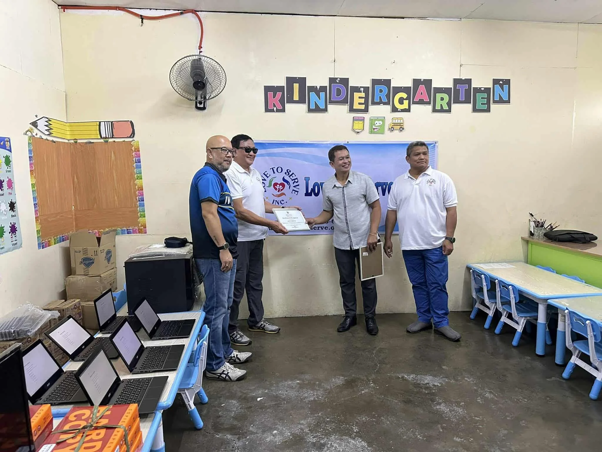 Four men standing in a kindergarten classroom with laptops and boxes on tables, one man receiving a certificate from another, a banner and colorful decorations in the background.