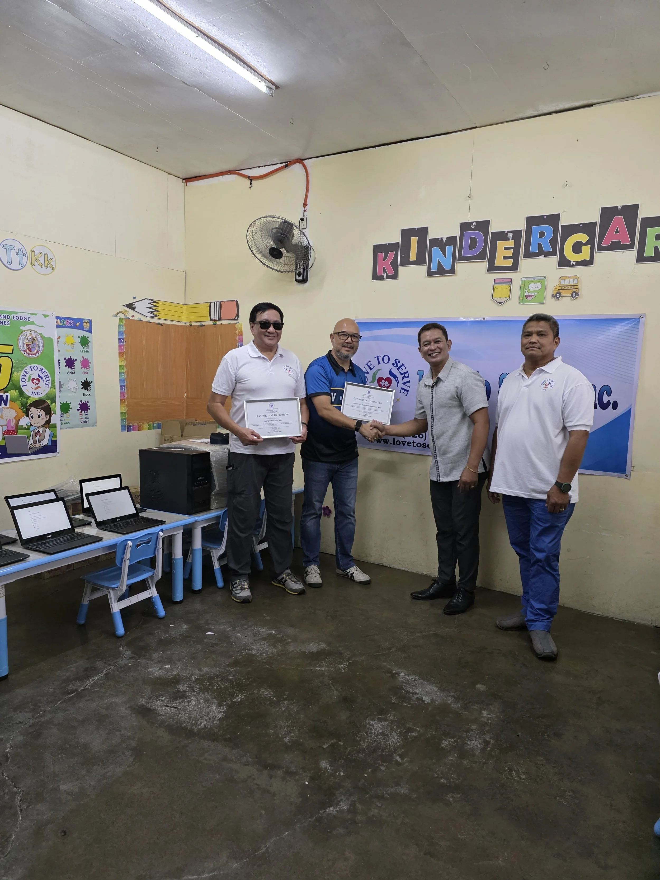 Four men in a classroom, with two holding certificates and shaking hands, in front of a banner with a heart and the words "Love to Serve." The classroom has computers and children's artwork on the walls, and a colorful "Kindergarten" sign.