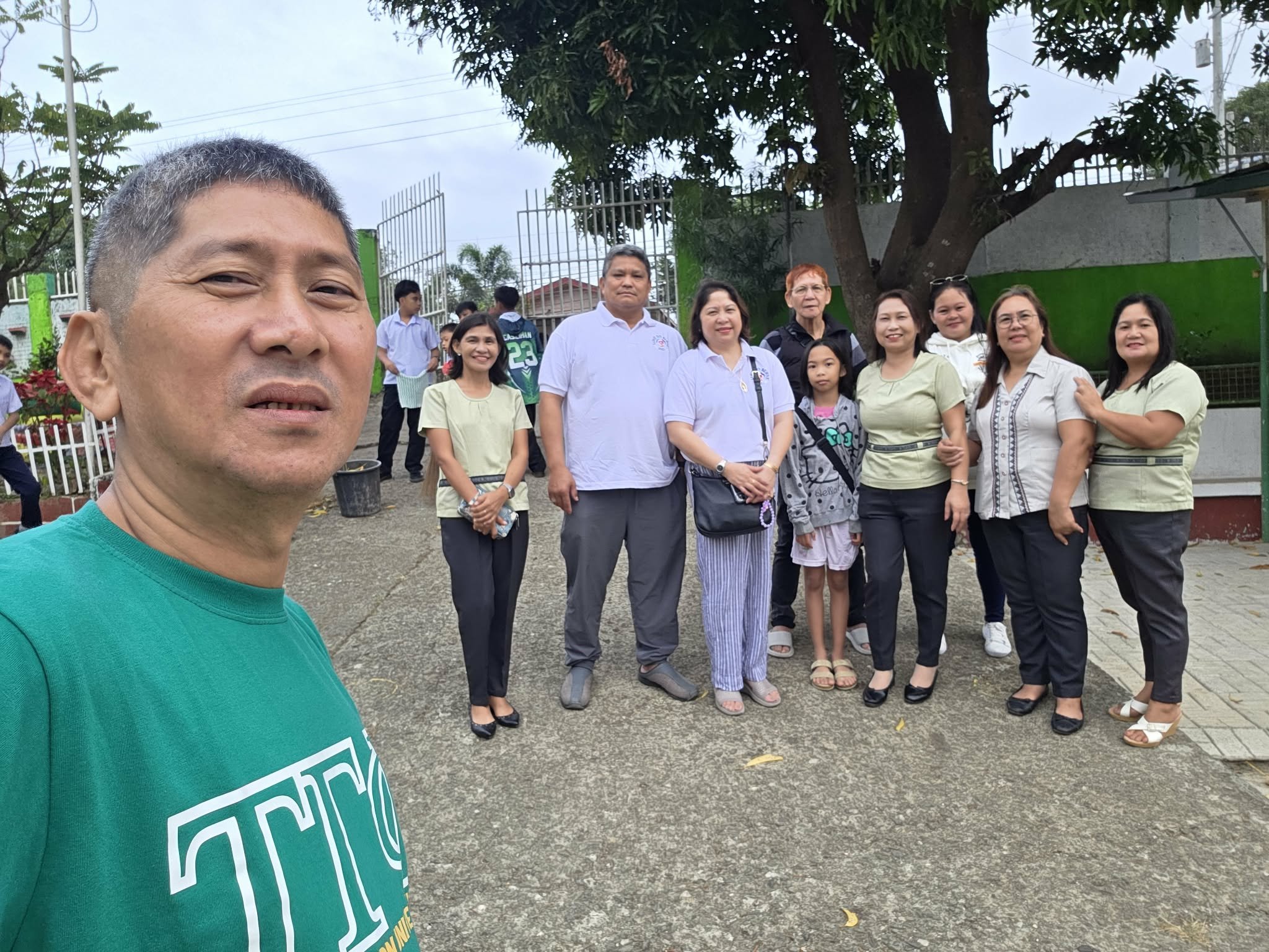A group of people posing outdoors in front of a fence and trees, with a man taking a selfie in the foreground.