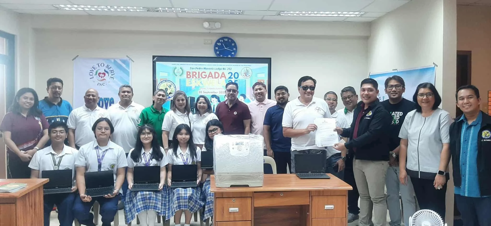 Group photo of adults and students in a classroom or meeting room during an event, with a banner and a screen displaying text in the background, some people holding documents, and laptops on the table in front.