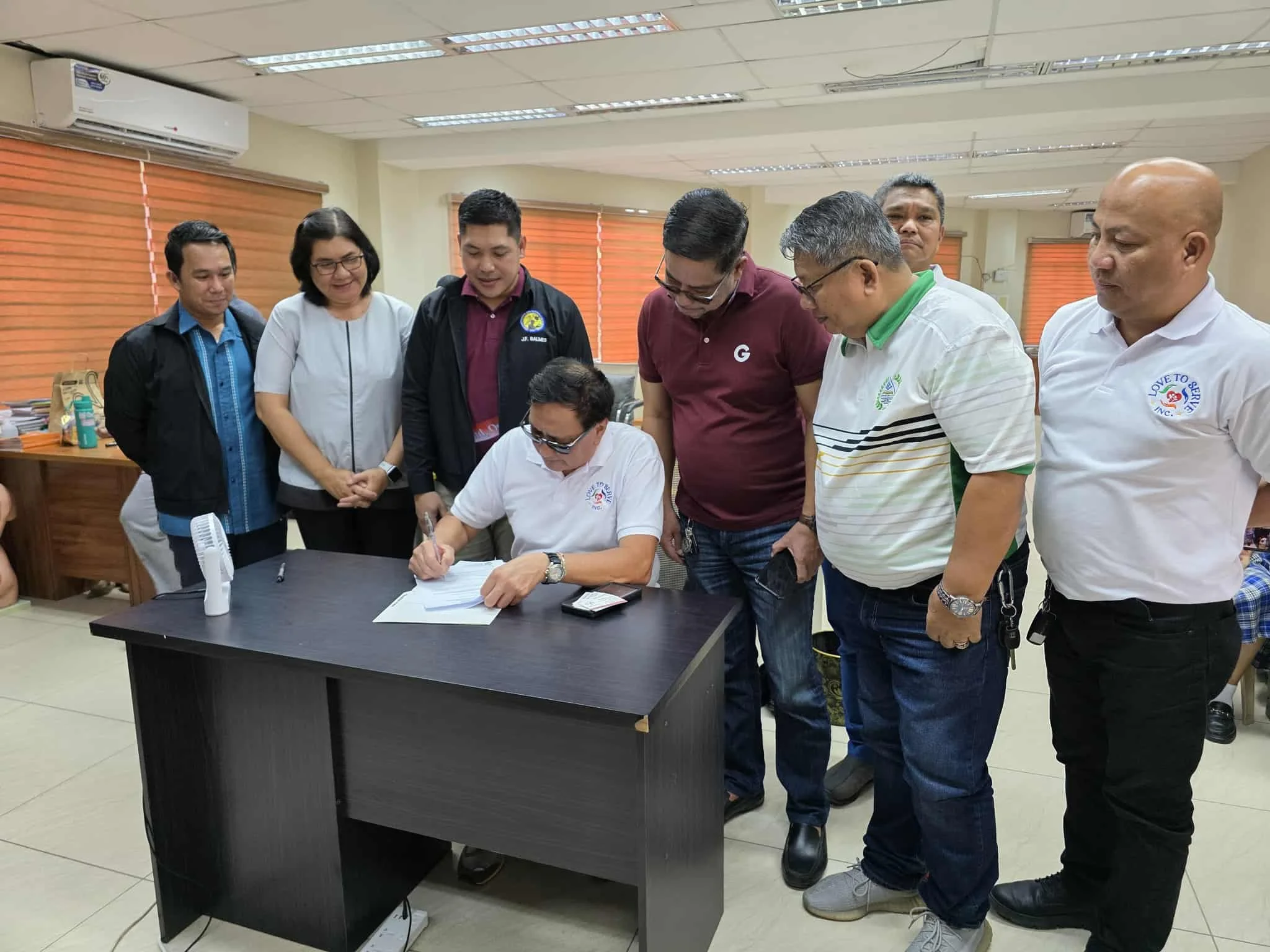 A man in white shirt is seated at a desk, signing a document, while seven other people stand around him, observing. The group includes men and women dressed in casual and business casual attire in an office setting with orange window blinds.